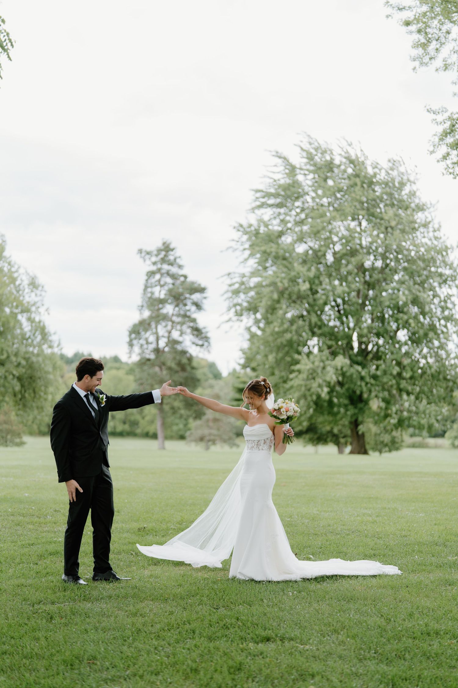 A couple in wedding attire dances and twirls together on a grassy field surrounded by trees.