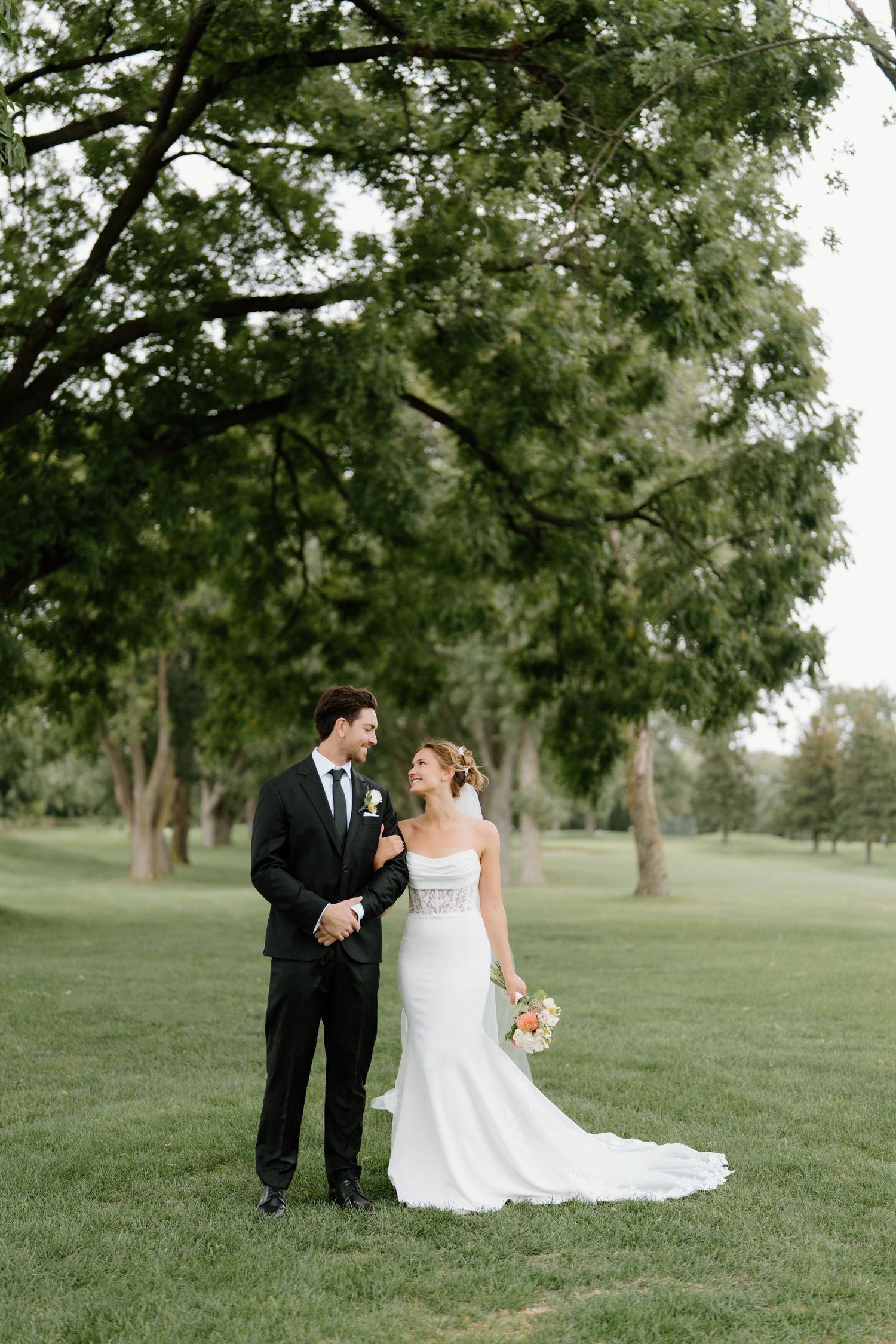 A wedding couple stands together on a beautiful lawn surrounded by tall trees.