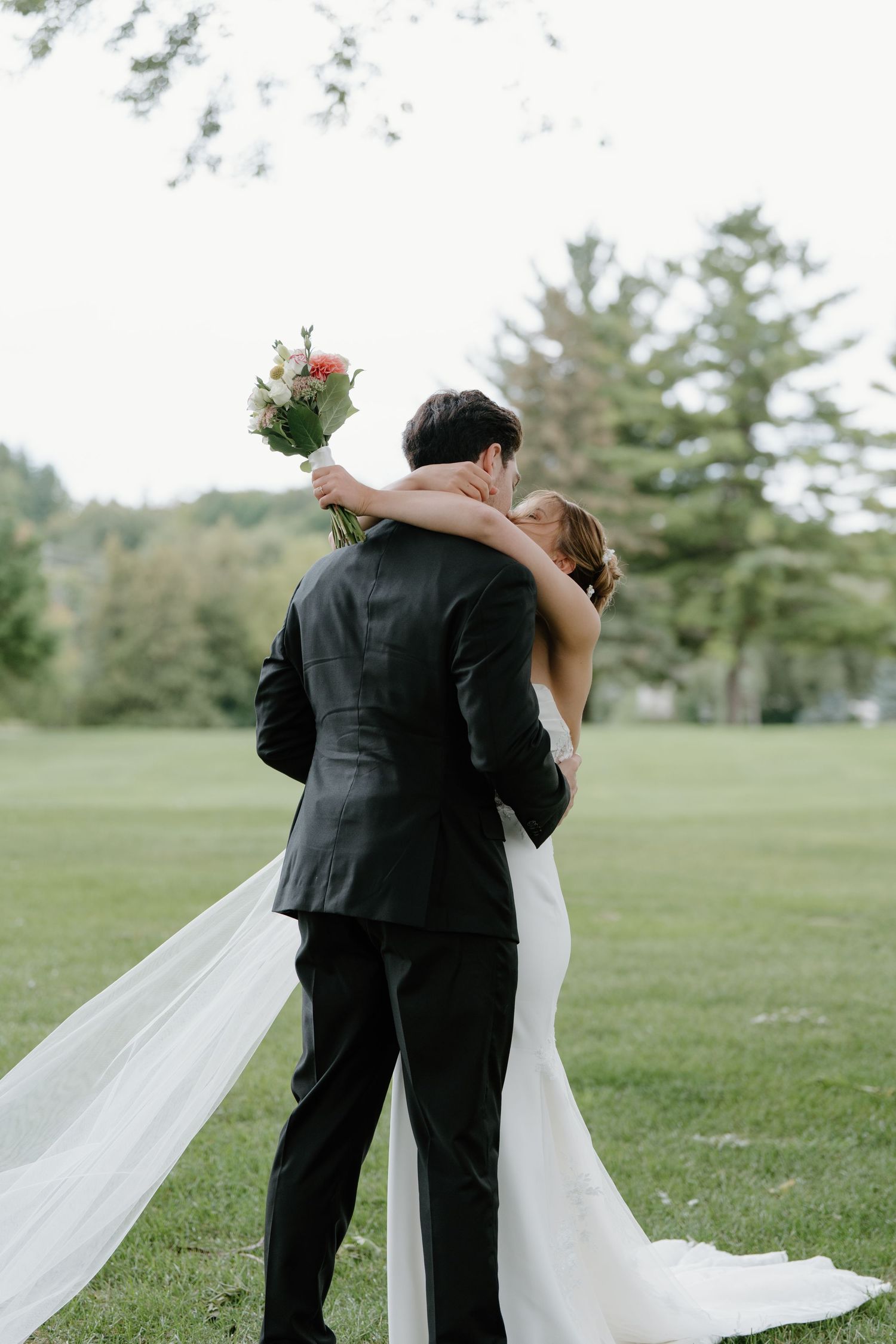A romantic wedding couple embraces in a lush green field as the bride's white veil flows gracefully in the breeze.