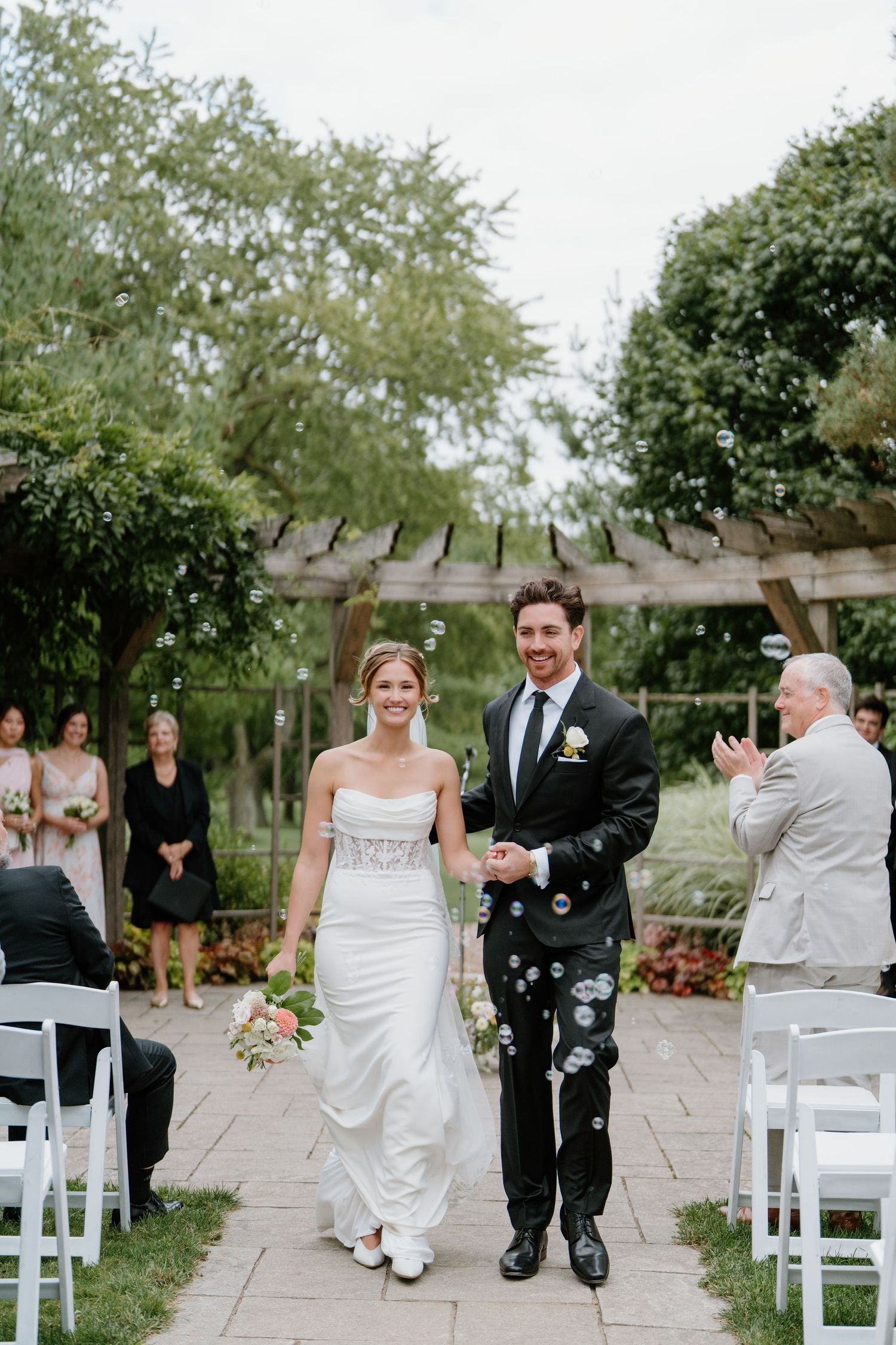 Newlyweds walk down the aisle together while guests celebrate with confetti at an outdoor garden wedding ceremony.