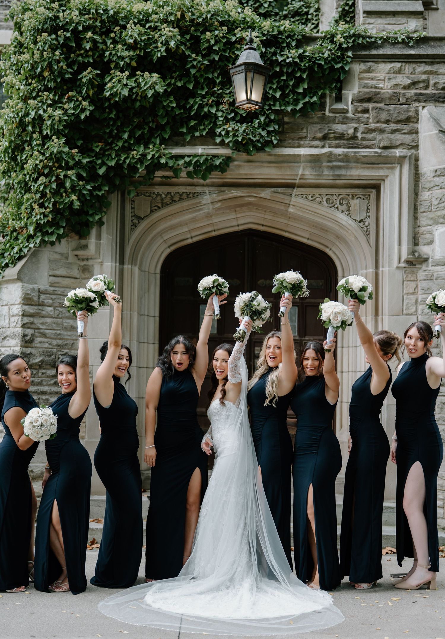 Wedding party in black attire stands in line against stone building while holding white bouquets aloft in celebration.
