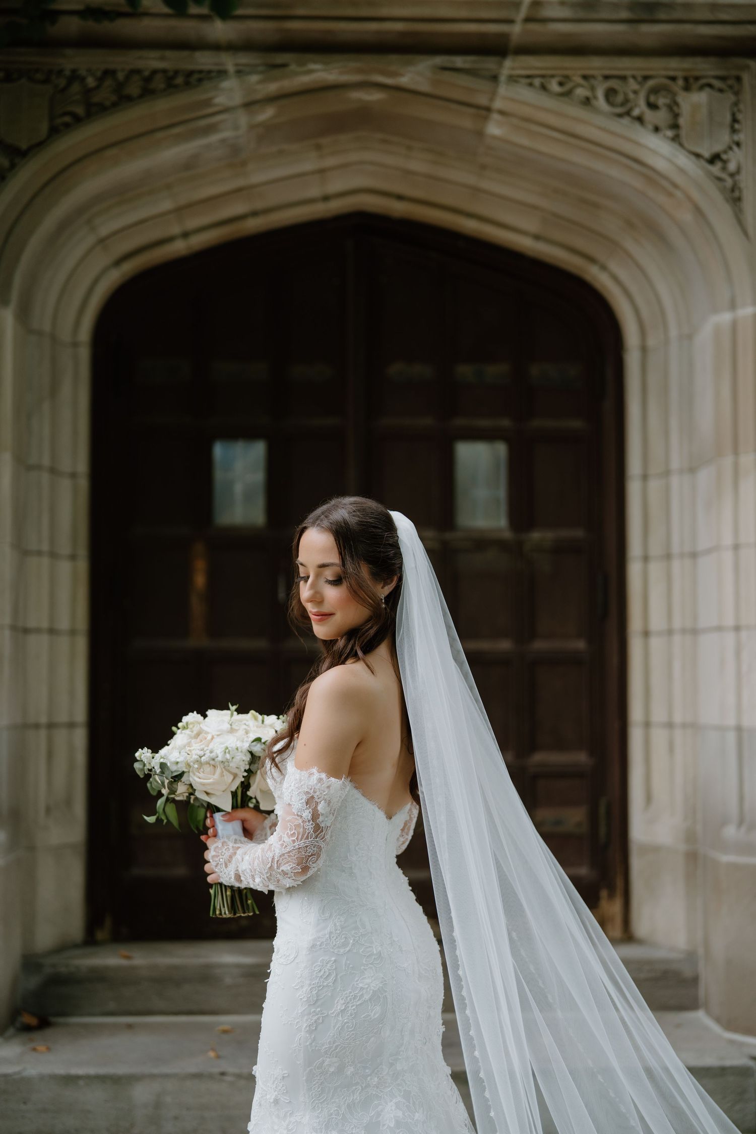 A bride with a dramatic cathedral veil holds a white bouquet against a gothic stone archway.
