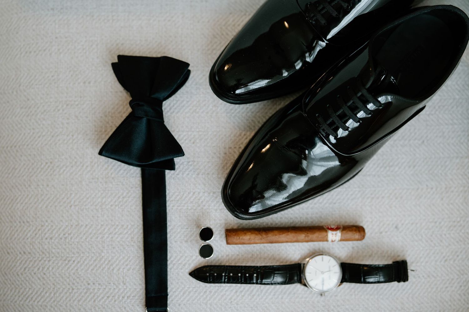 A collection of formal men's accessories including black bow ties, dress shoes, and a cigar arranged on a light surface.