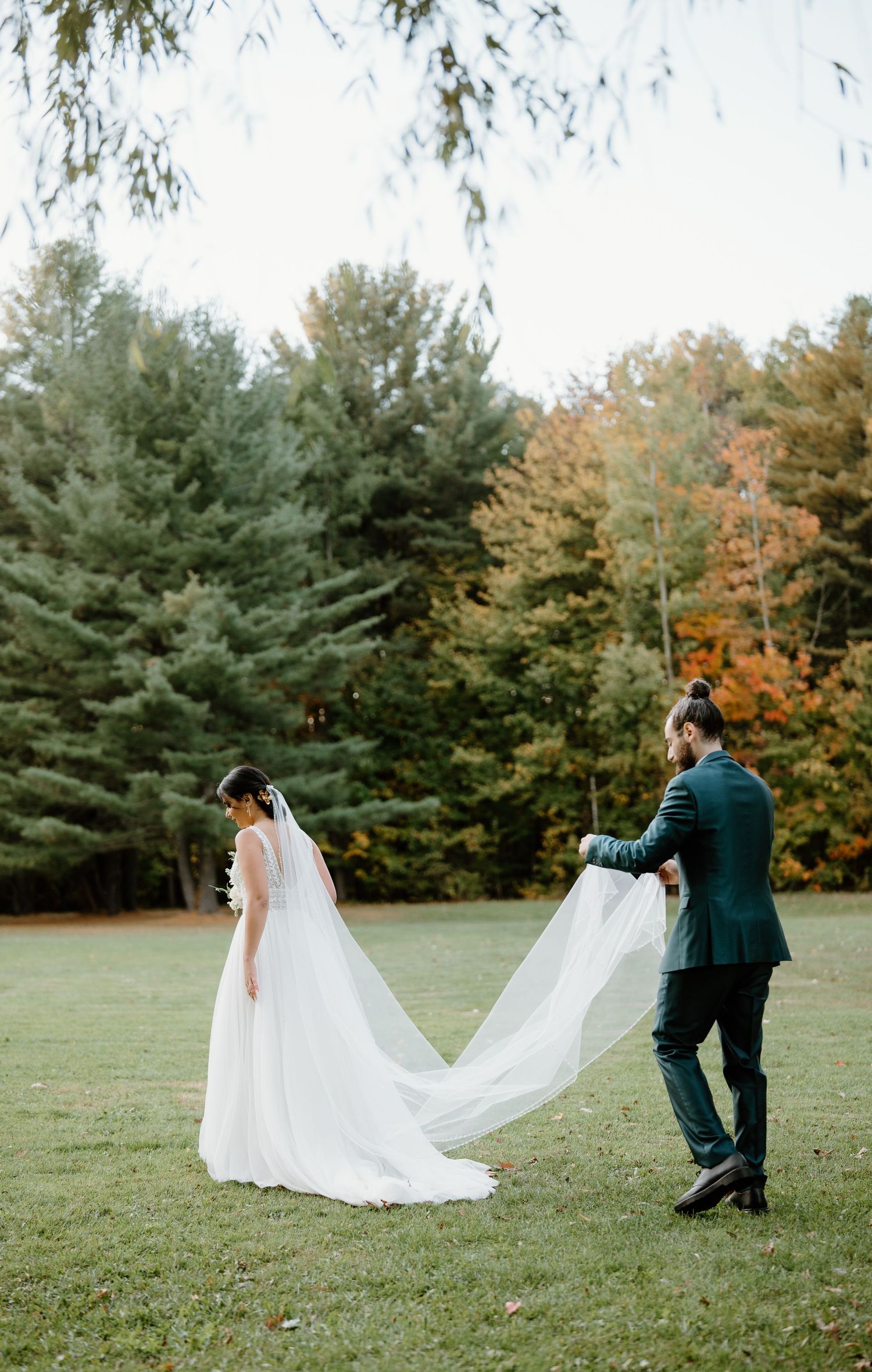 Couple in wedding attire walking through autumn field with flowing veil trailing behind.