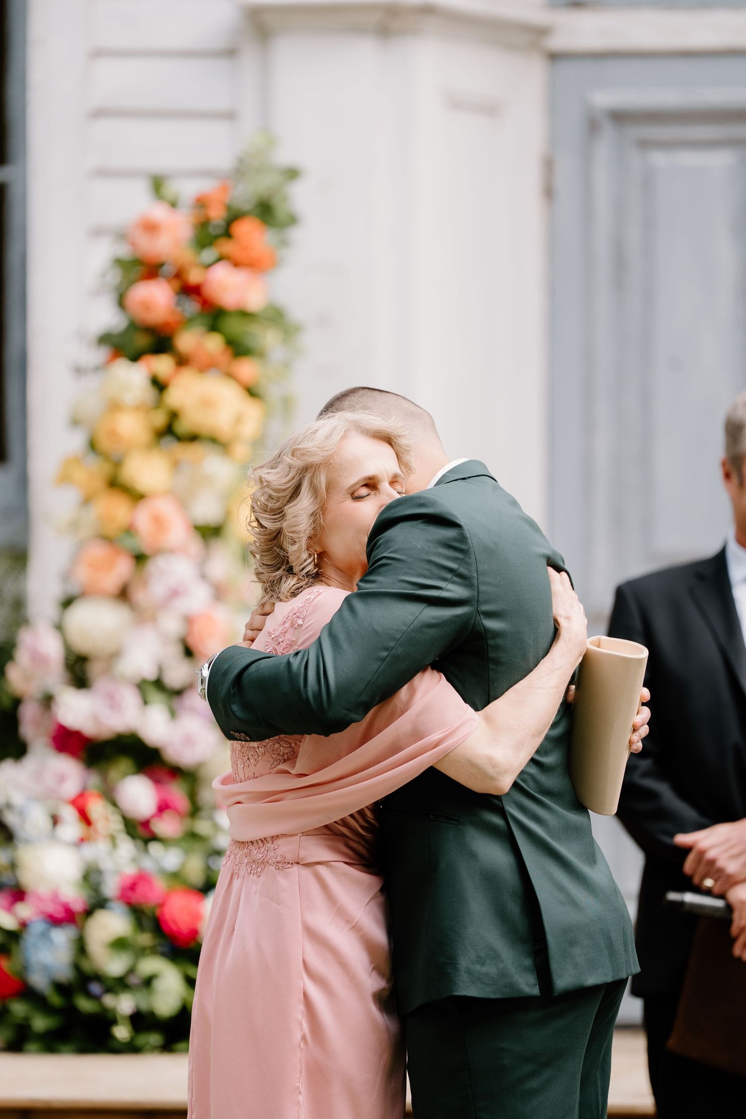 A couple embraces during a wedding ceremony with peach and coral floral arrangements in the background.