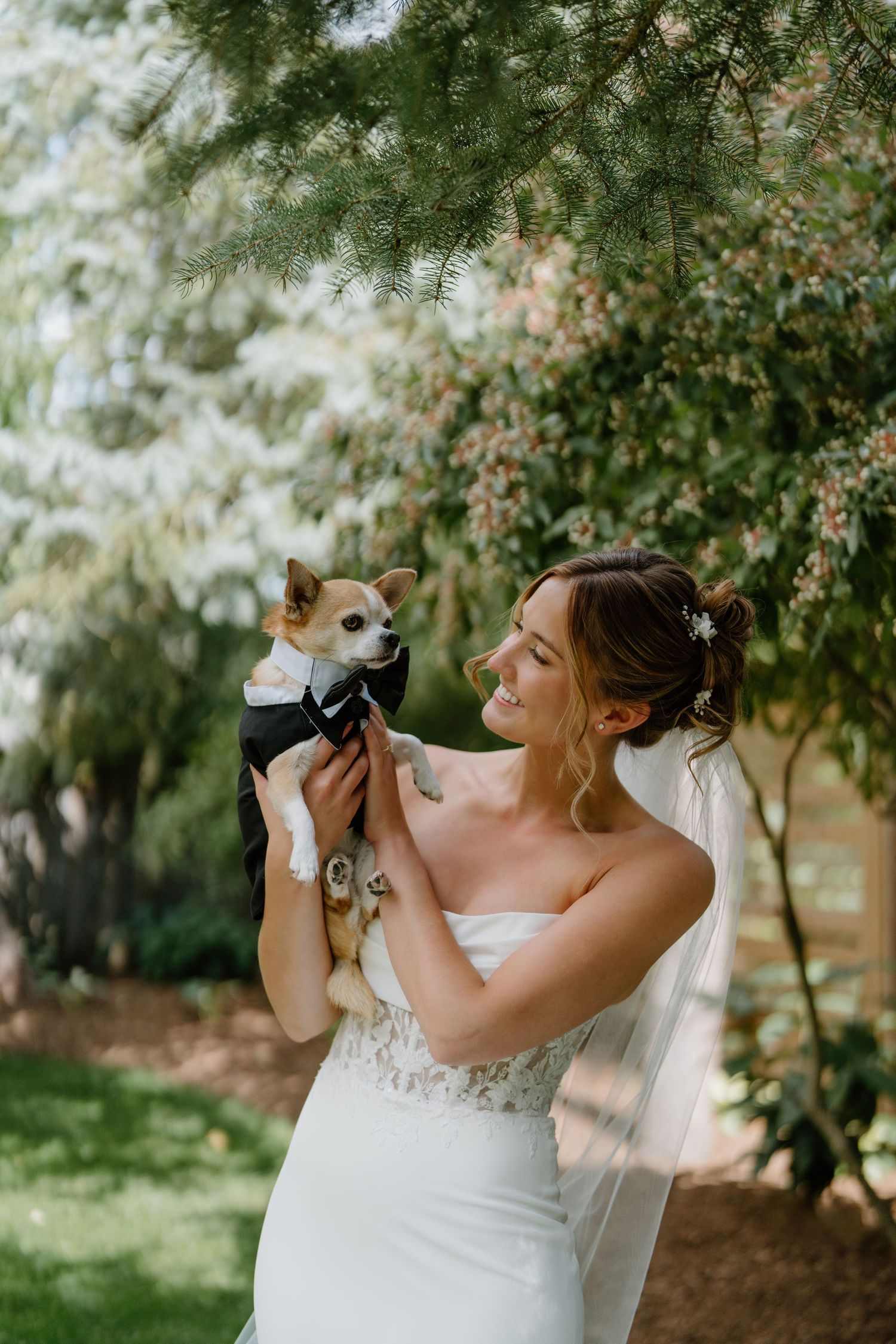 A bride in a white lace wedding dress takes a celebratory drink from a bottle in an outdoor garden setting.