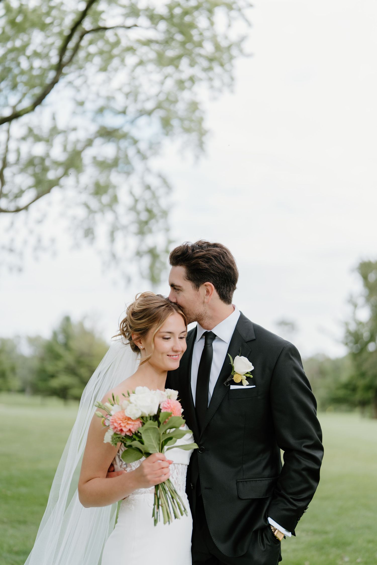 A romantic wedding couple shares intimate moments outdoors under trees on their special day.