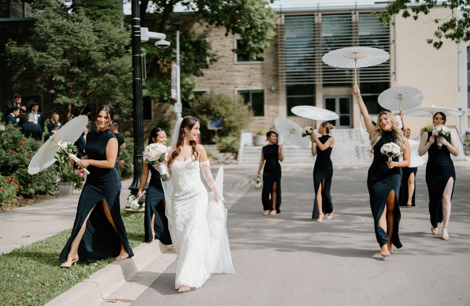 Wedding party in black and white dresses walks along a stone path carrying parasols.