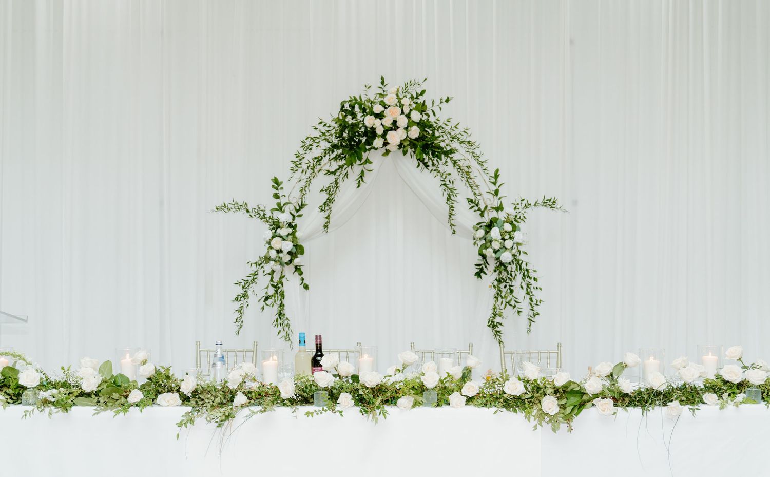 A delicate floral wedding arch with white flowers and greenery against a light gray wall.