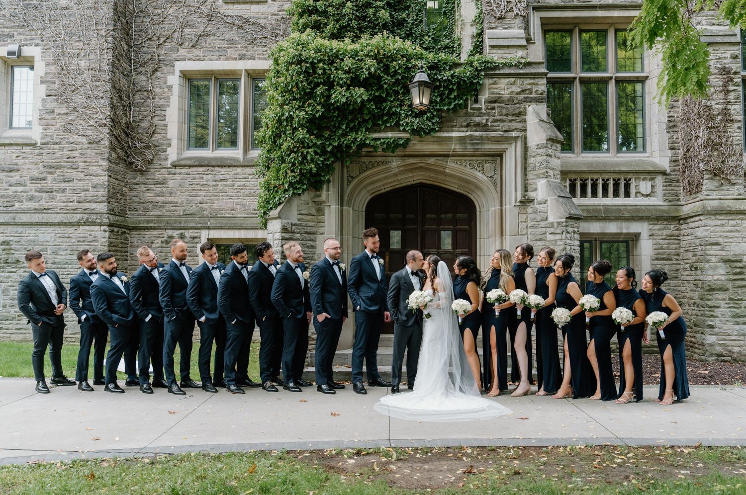 Wedding party in navy suits and black dresses lined up against stone building exterior.