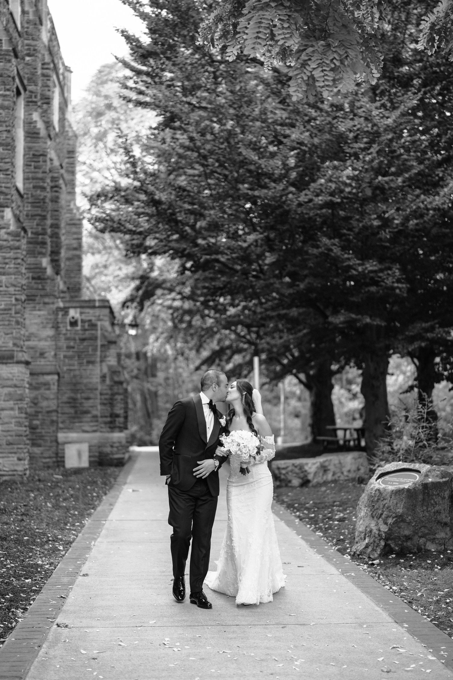Wedding couple walking together on a tree-lined path outside a Gothic stone building.