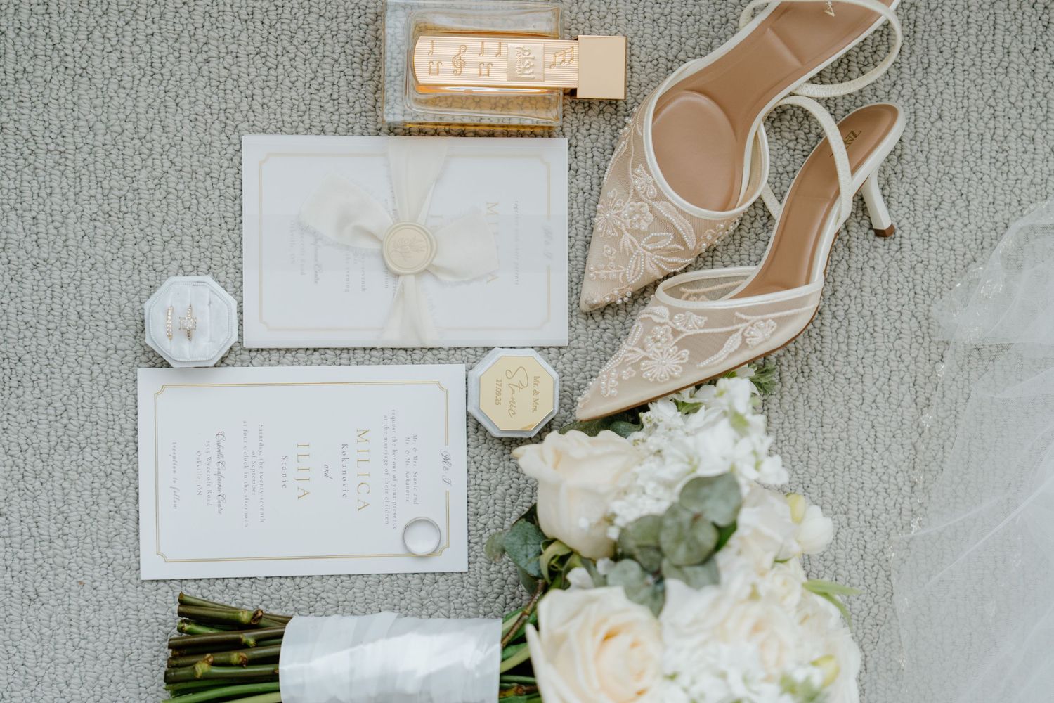 Wedding accessories including bridal shoes, invitations, rings, and white roses arranged on a light gray surface.