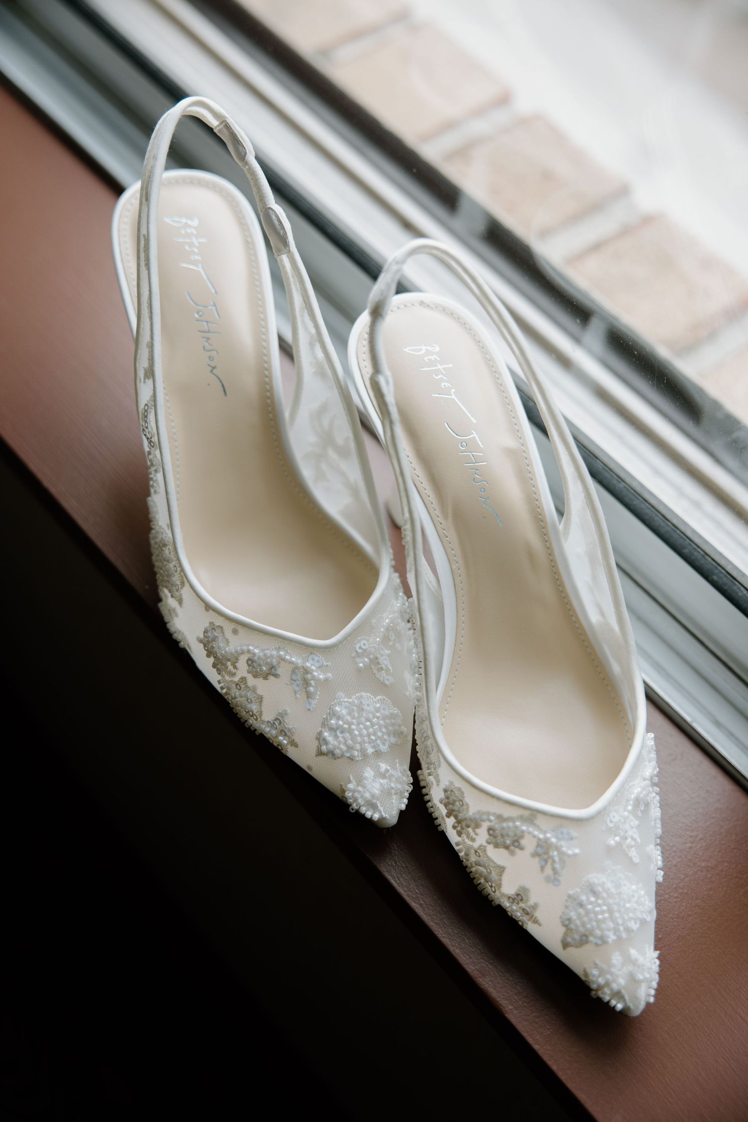Elegant white lace bridal slingback heels with delicate beading details sit on a window ledge.