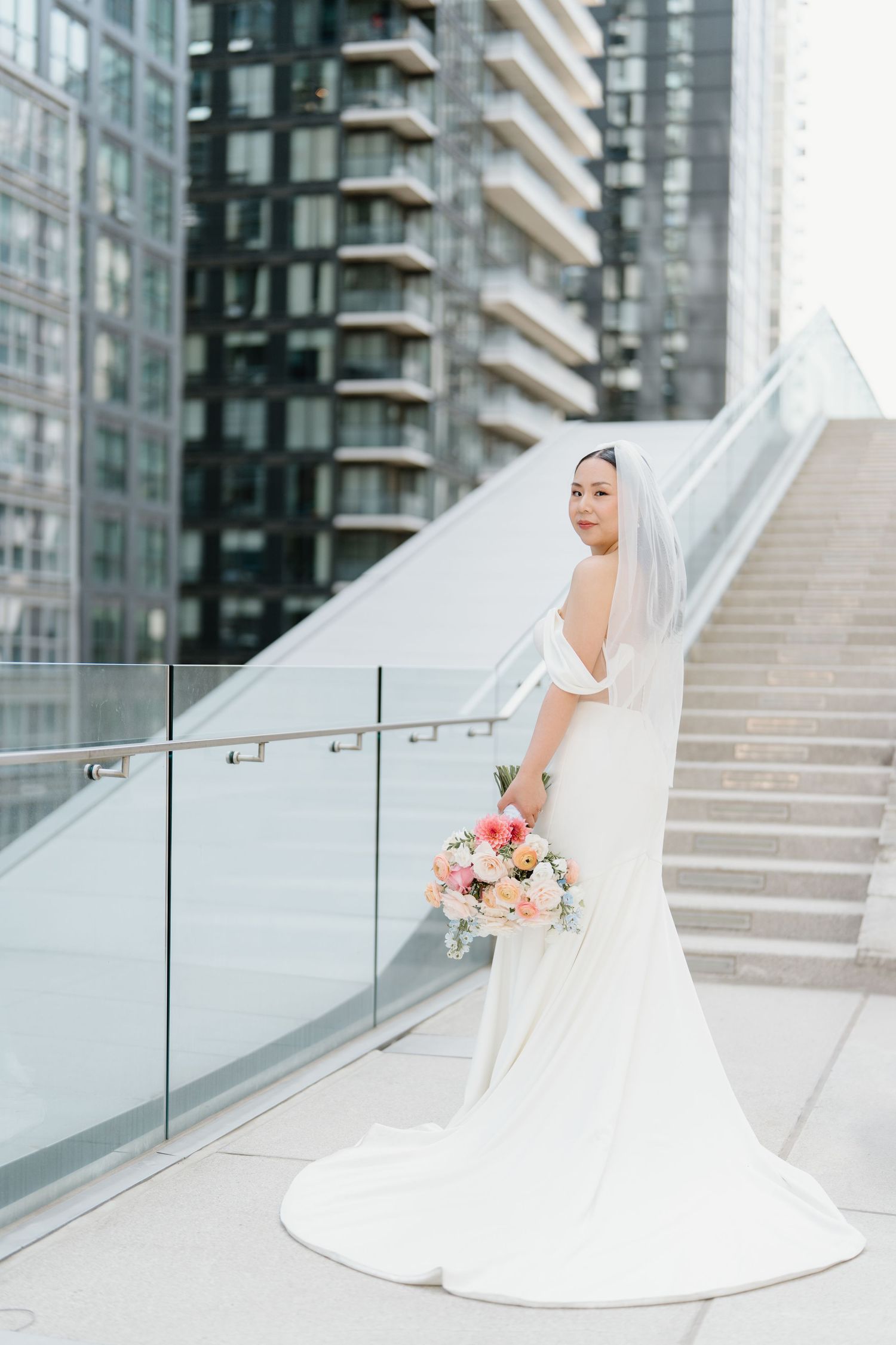 A bride in a flowing white gown and veil poses with a pink and peach bouquet on modern urban stairs with glass railings.