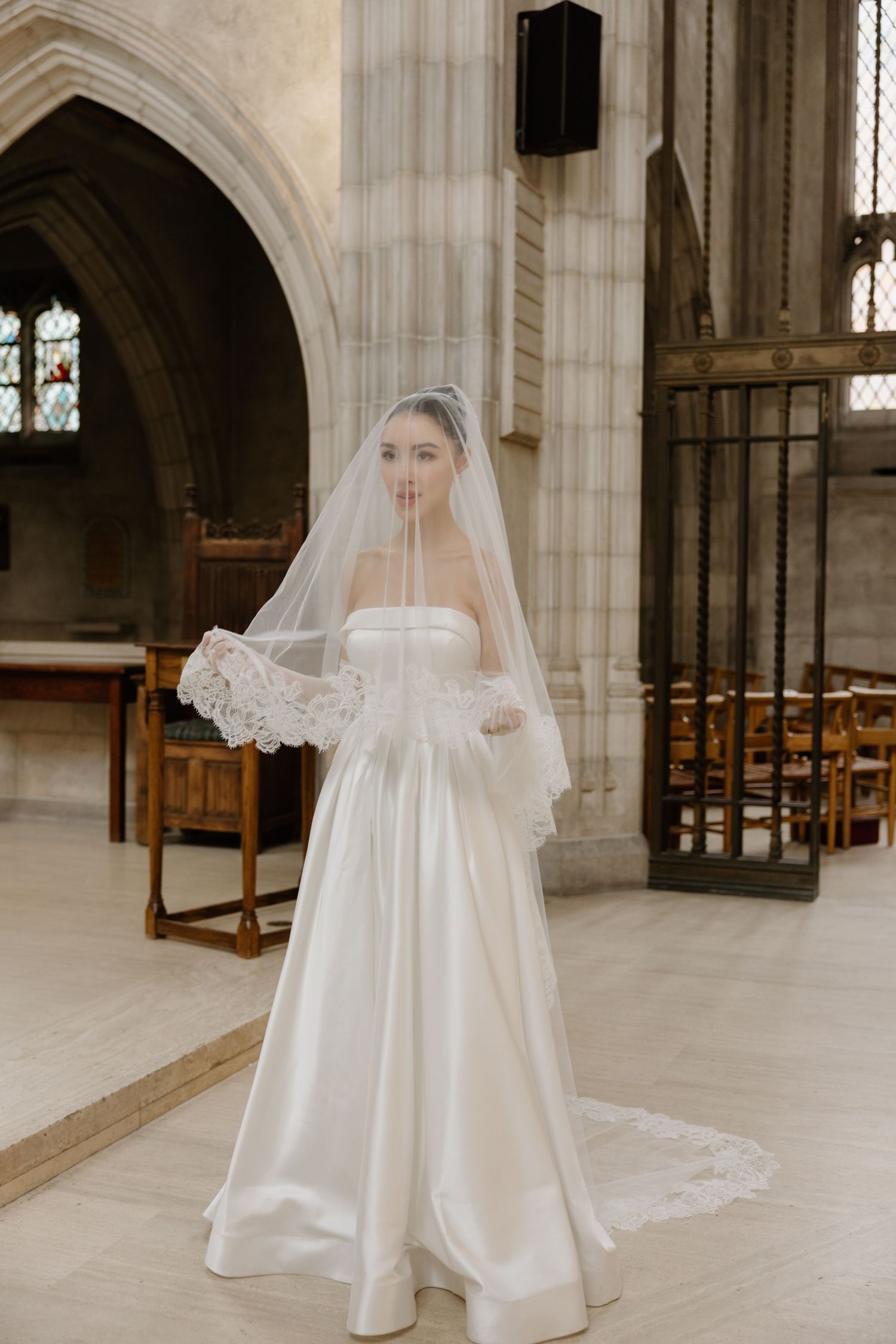 A bride wearing an elegant white satin wedding gown with a long lace-trimmed veil stands in a gothic church interior.