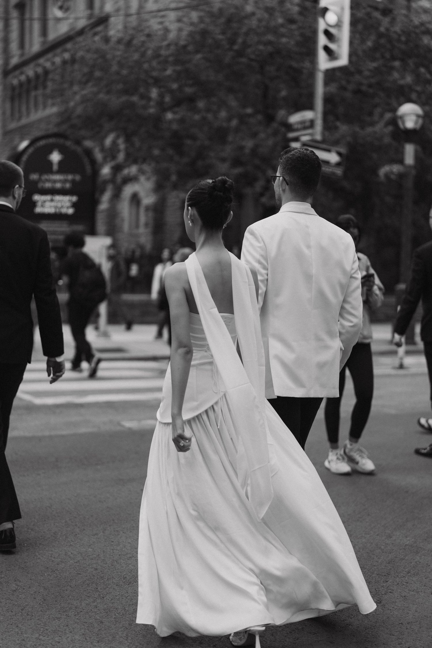 A couple in elegant white attire walks down a city street in a black and white photograph.