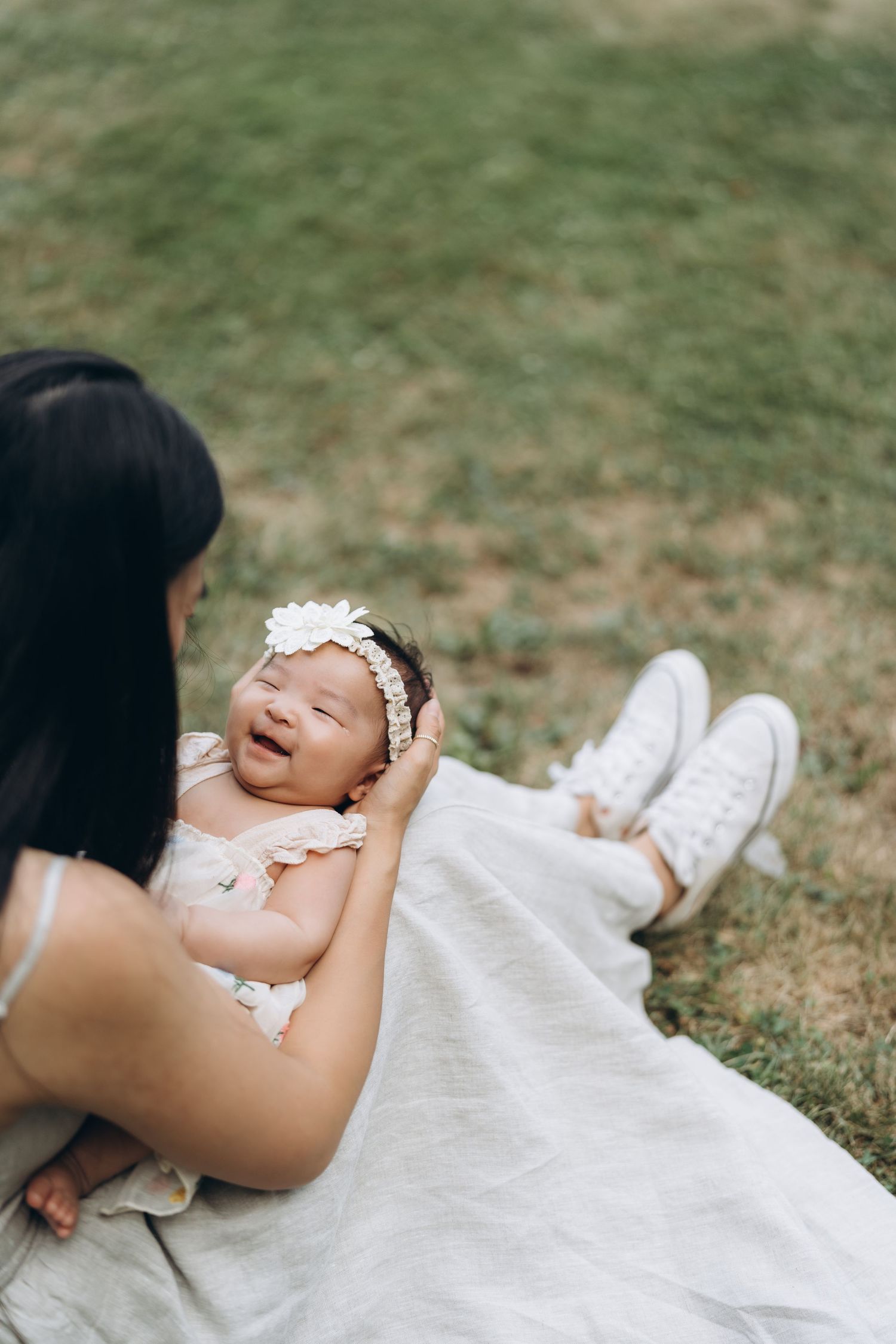 A happy newborn baby wearing a white floral headband lies on a blanket while being cradled outdoors on grass.