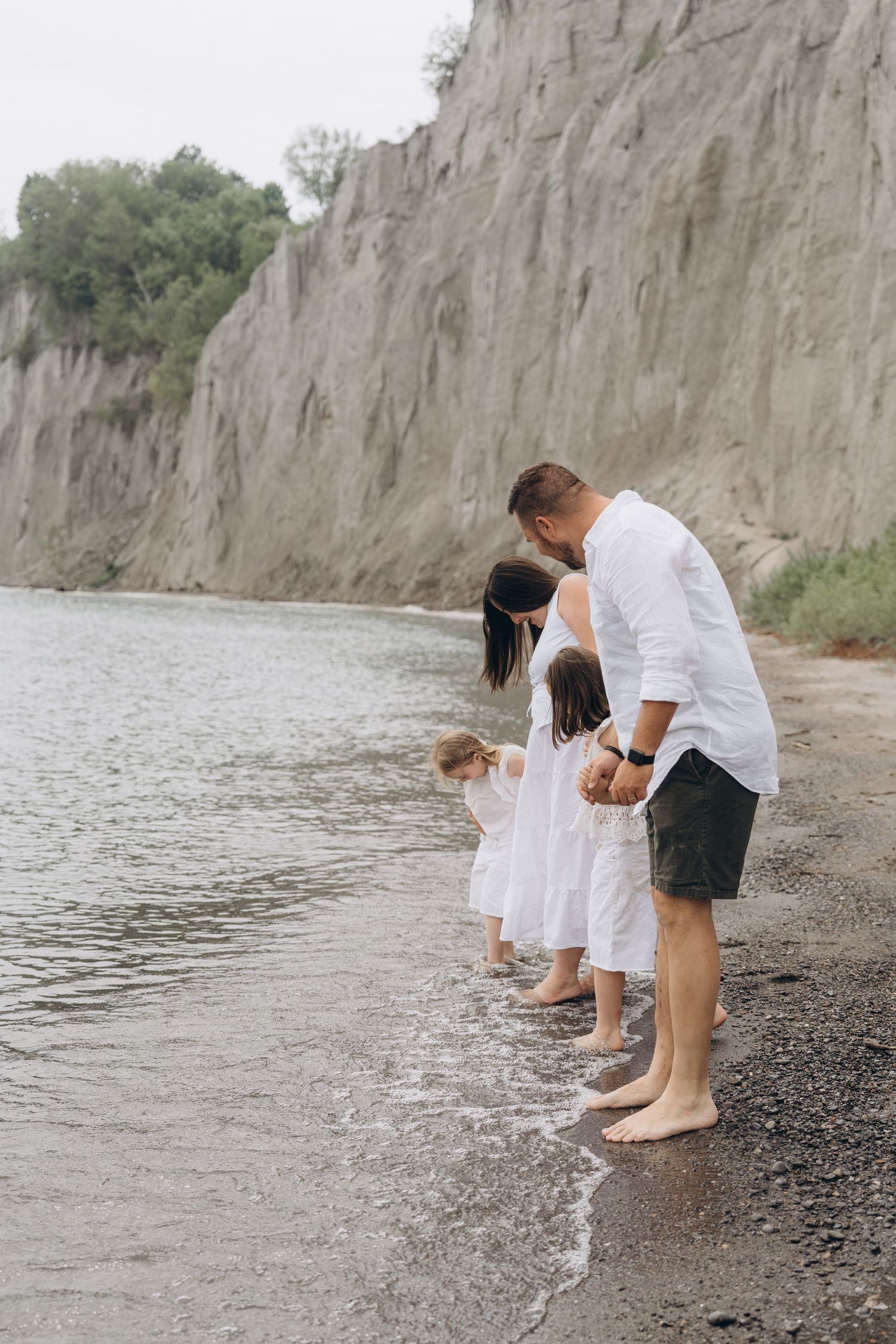 People in white outfits dipping their feet in water at a lakeside beach with tall cliffs in the background.