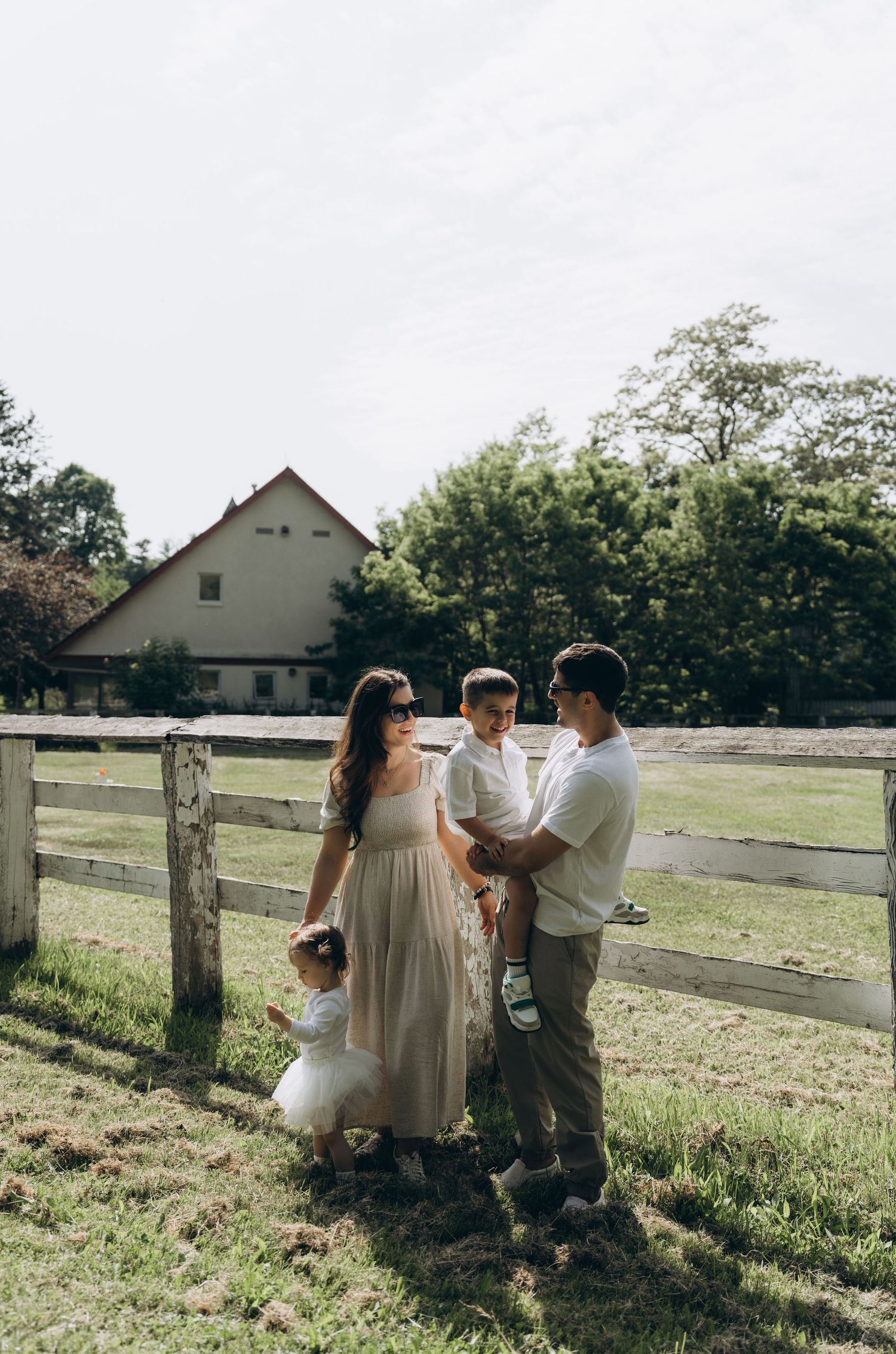 A rustic outdoor family photoshoot with people leaning against a wooden fence on a sunny day in a pastoral setting.