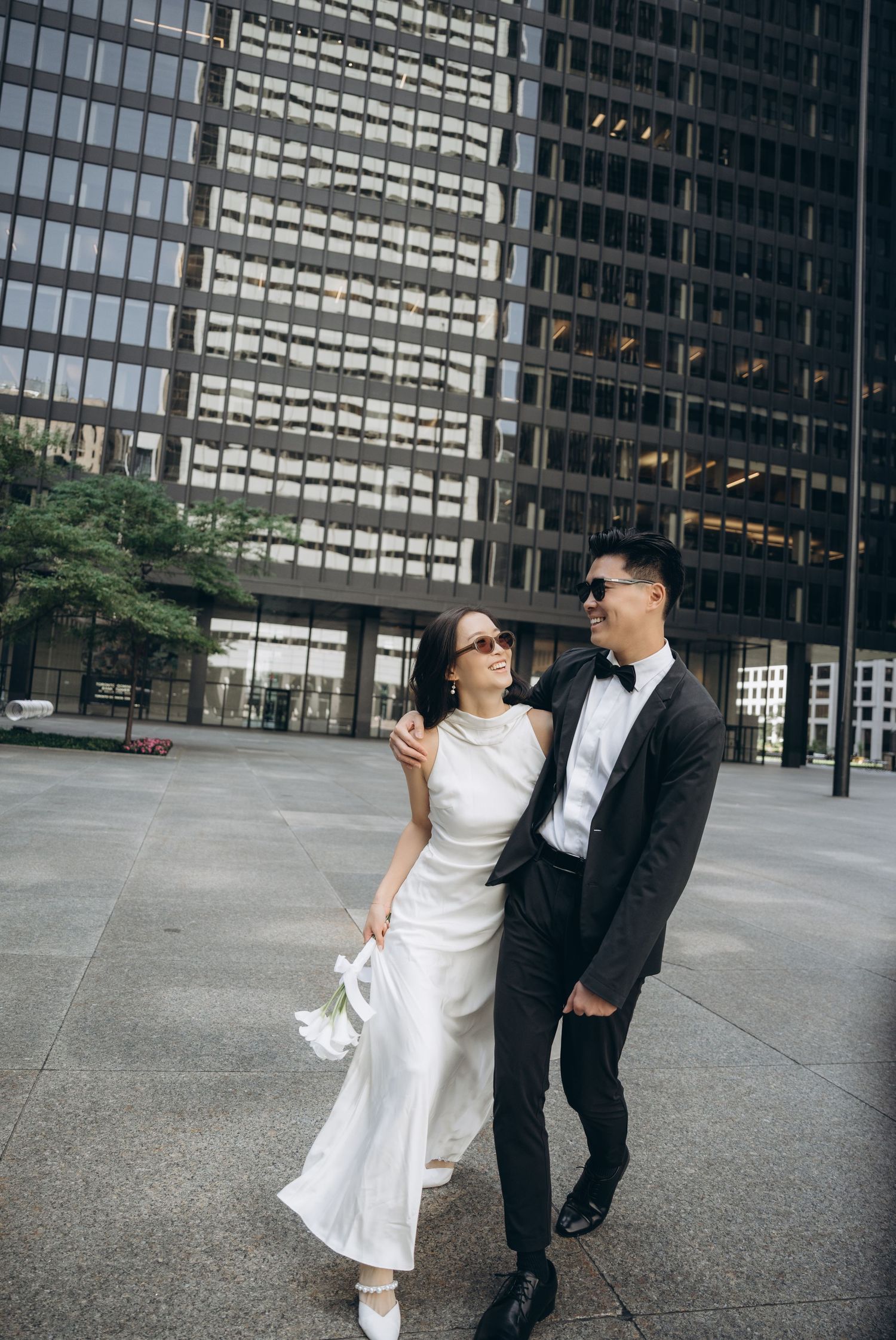 Couple in wedding attire dance and pose playfully in front of modern glass office building.