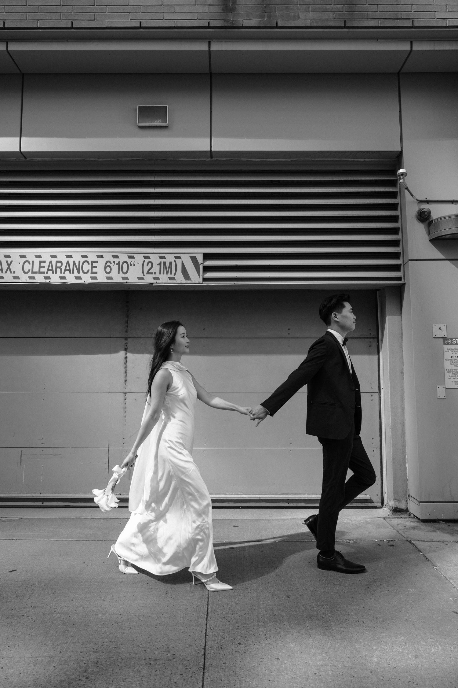 A bride and groom walking hand in hand under a city clearance sign in black and white photography.