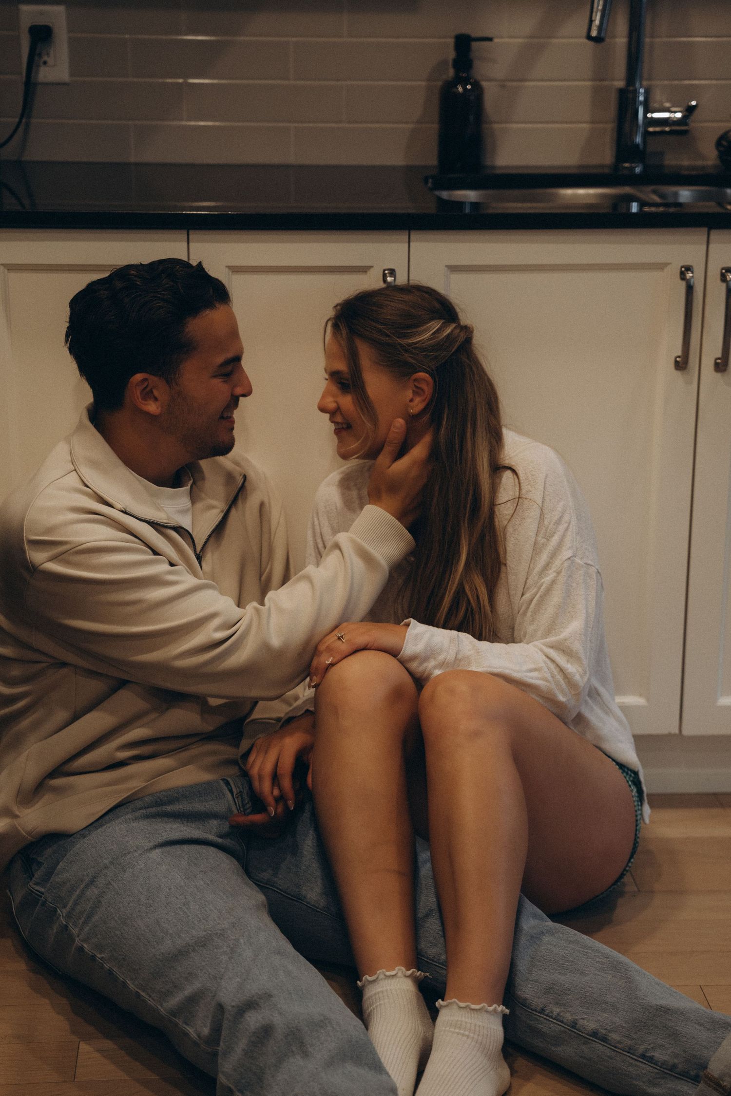 A couple in casual outfits sits together intimately on their kitchen floor in warm lighting.