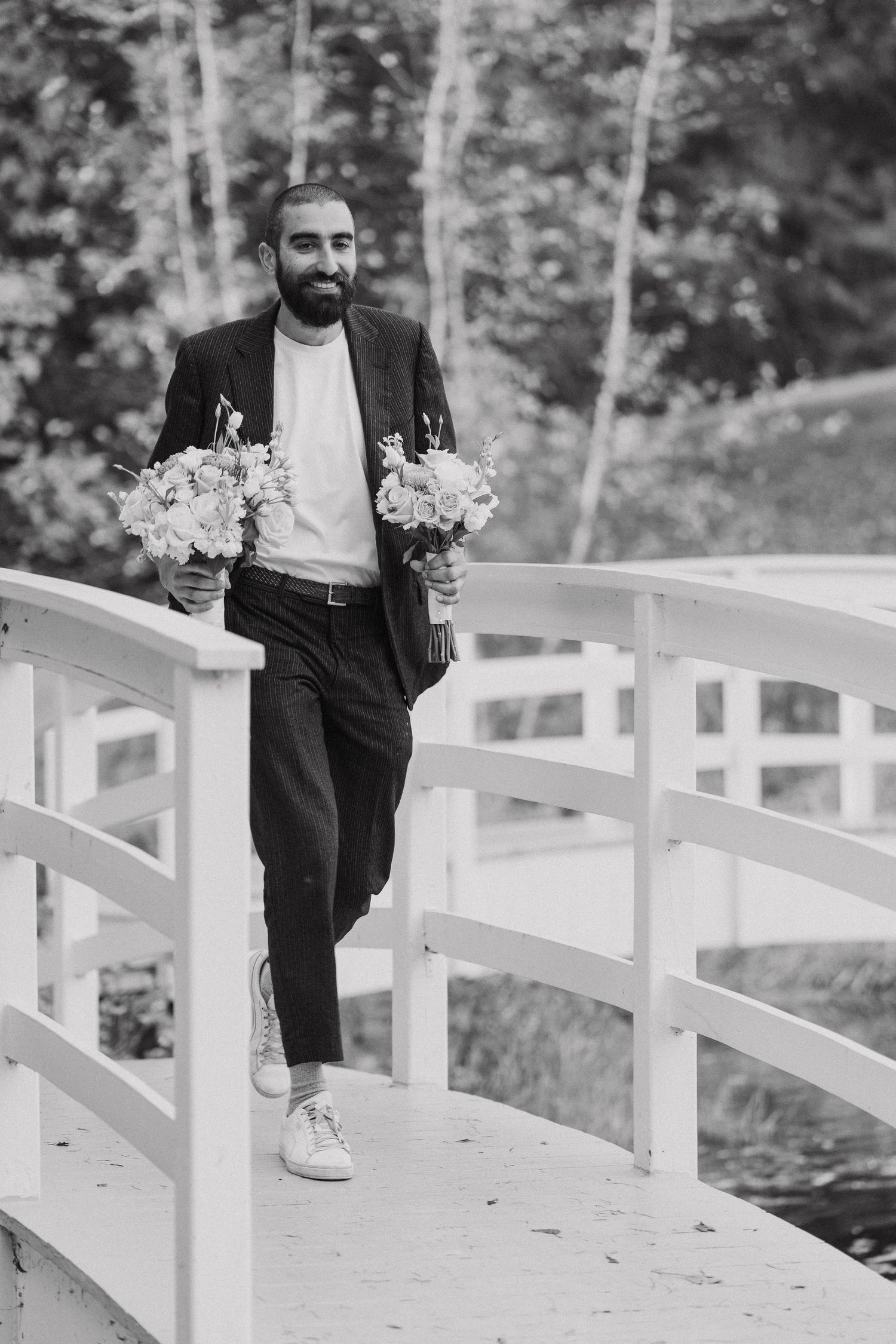 Man in dark suit carrying flower bouquets while walking on white bridge.