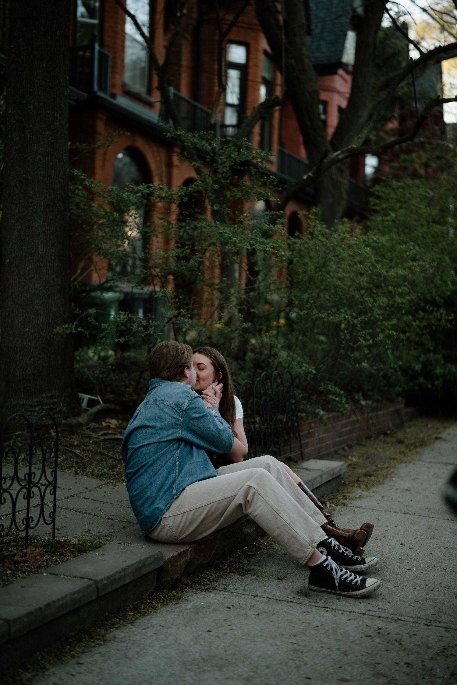 A couple shares an intimate moment sitting on a curb outside historic brownstone buildings.