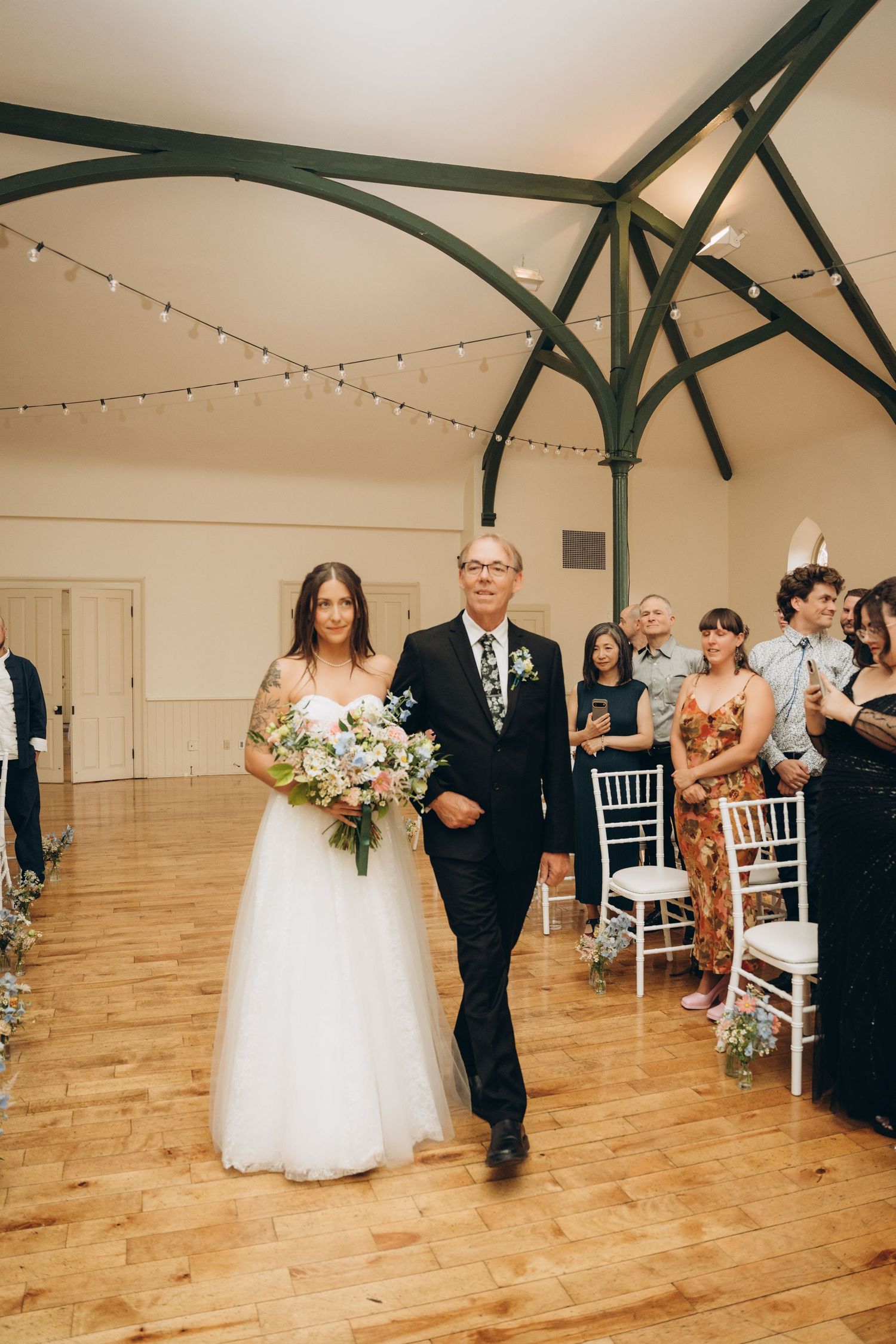 Bride in white gown walks down wooden-floored aisle with escort as guests watch from white chairs.