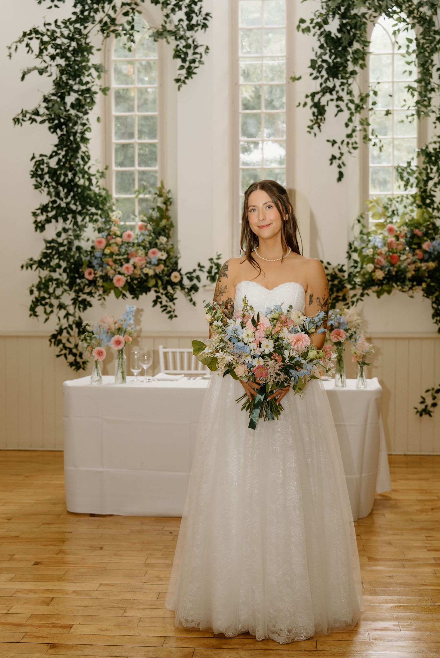 A bride in a white gown poses elegantly at a wedding reception venue decorated with greenery and floral arrangements.