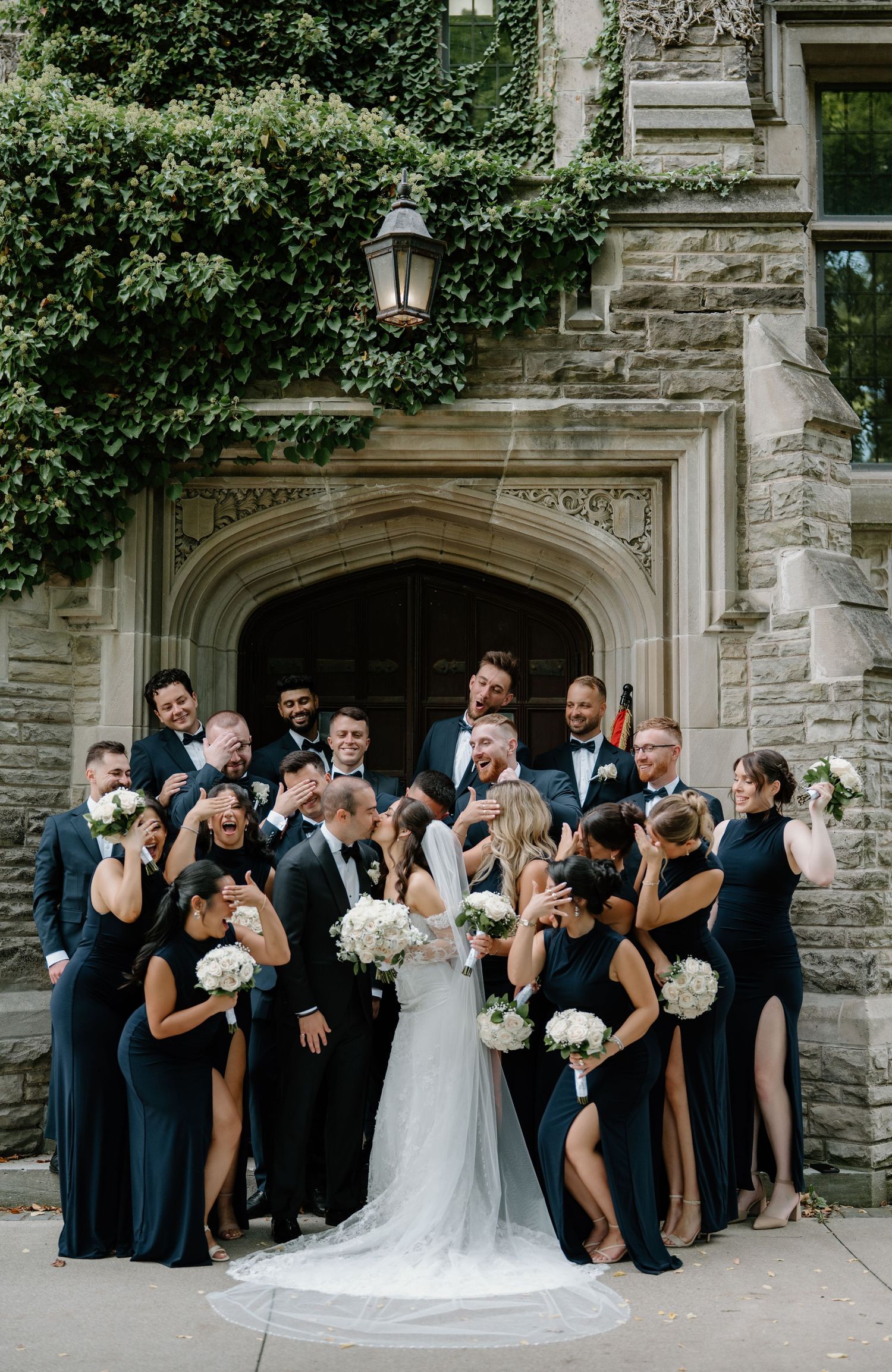 Wedding party in navy blue and white attire poses playfully in front of stone archway entrance.