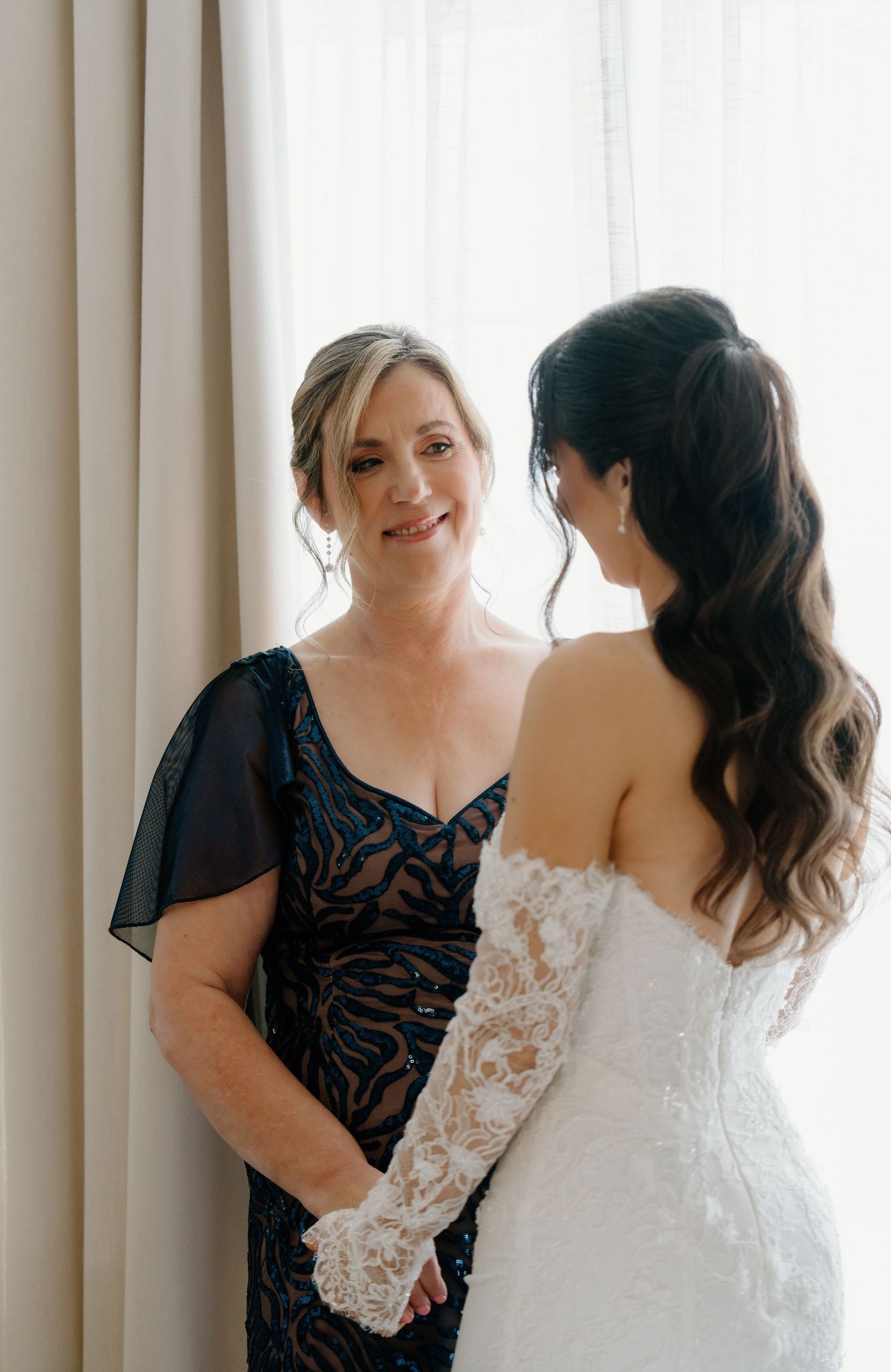 A bride in a white lace gown shares a joyful moment with someone wearing a black dress by a window.