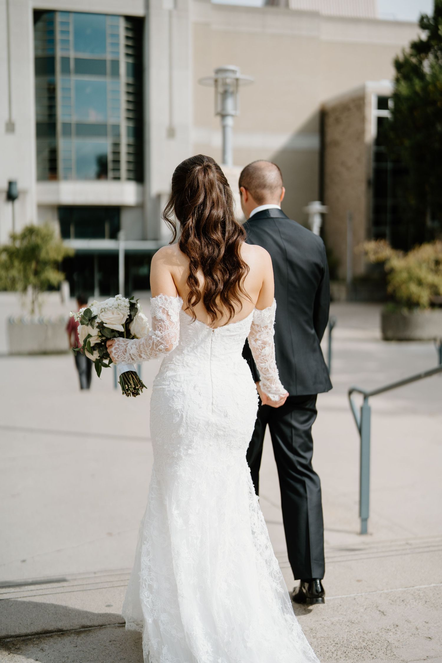 A bride and groom walk together on a modern building plaza with elegant wedding attire.