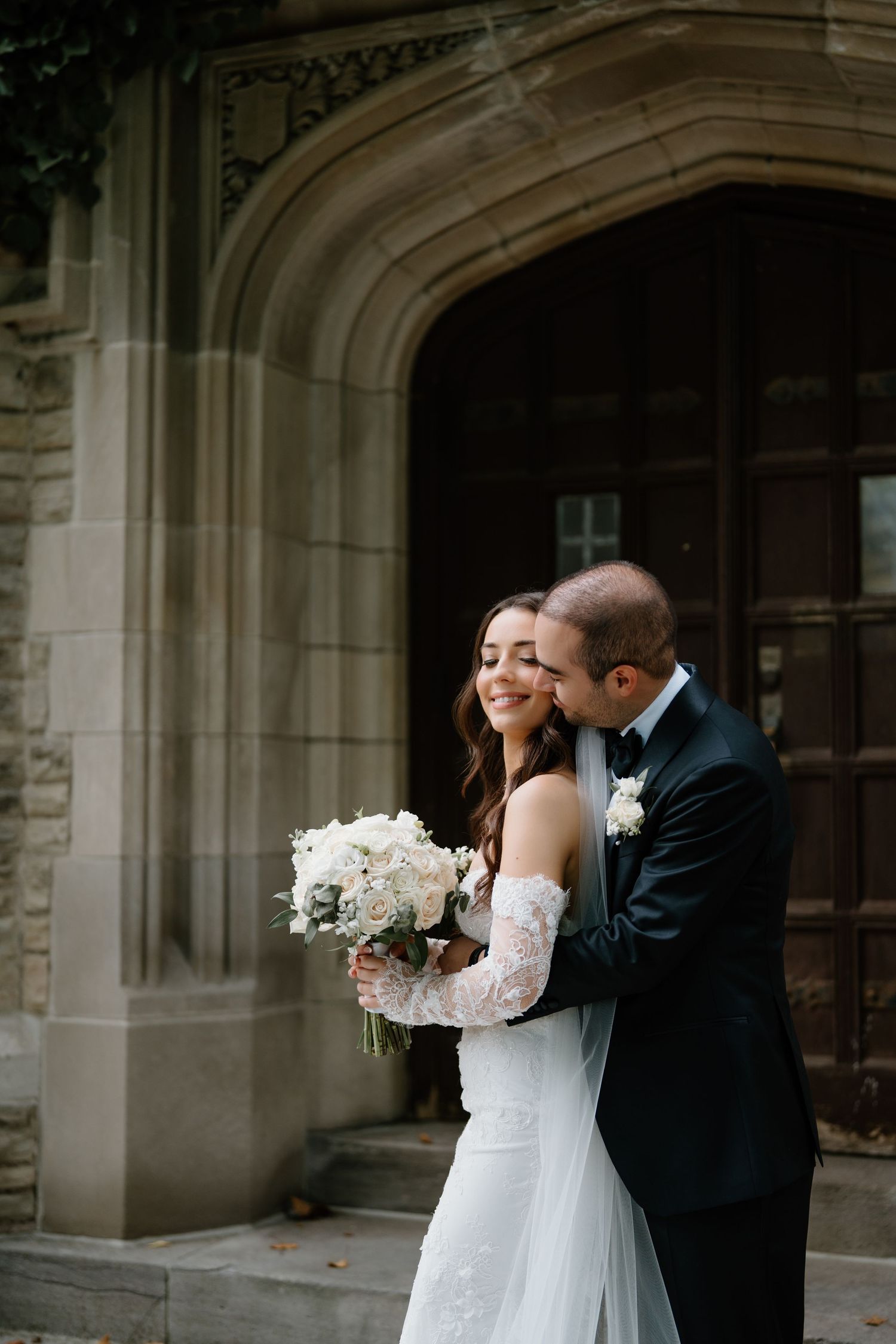 A romantic wedding moment captured in front of a stone archway with a couple sharing a tender embrace.