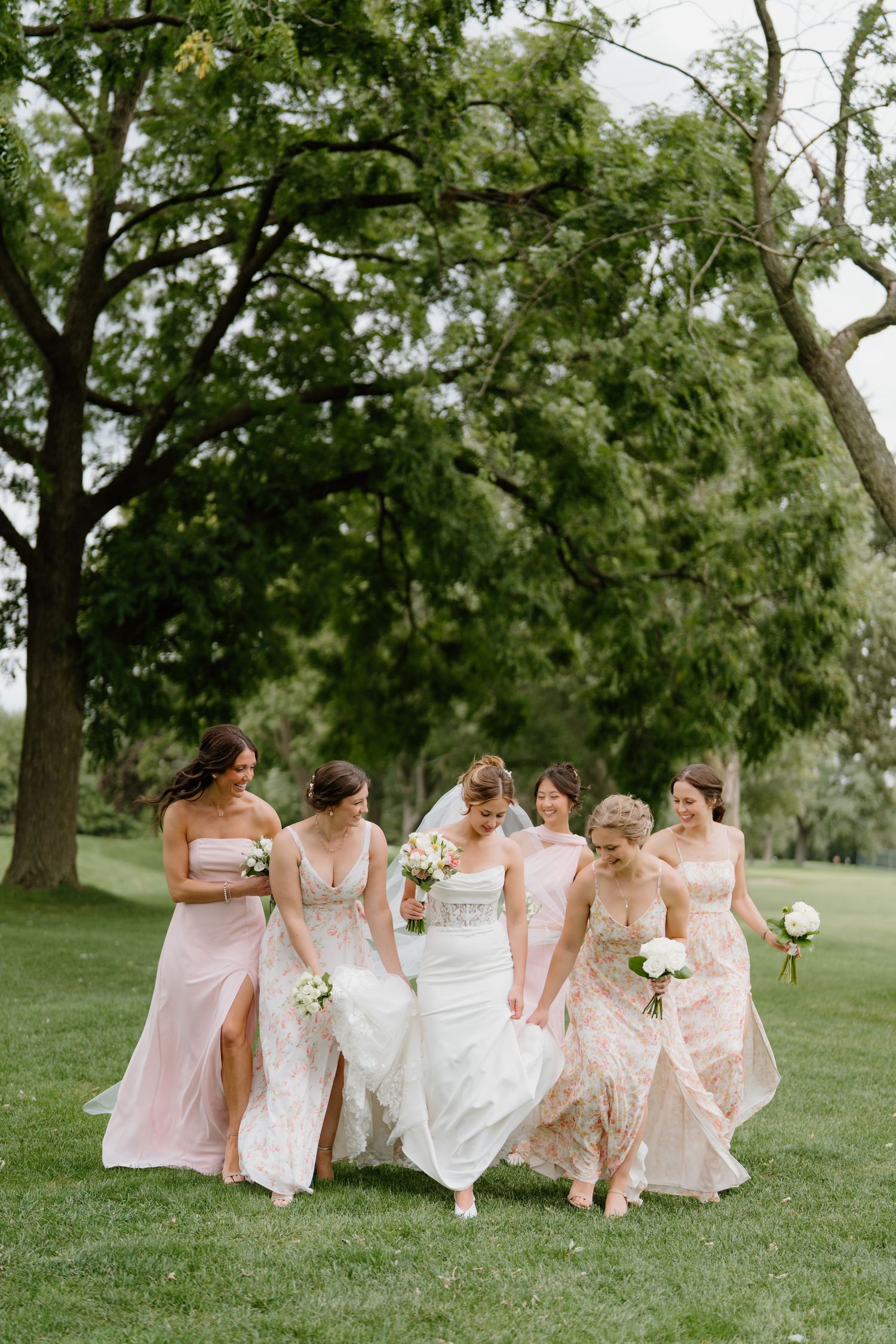 Bridal party in pink and white dresses runs playfully across a grassy lawn on a summer day.