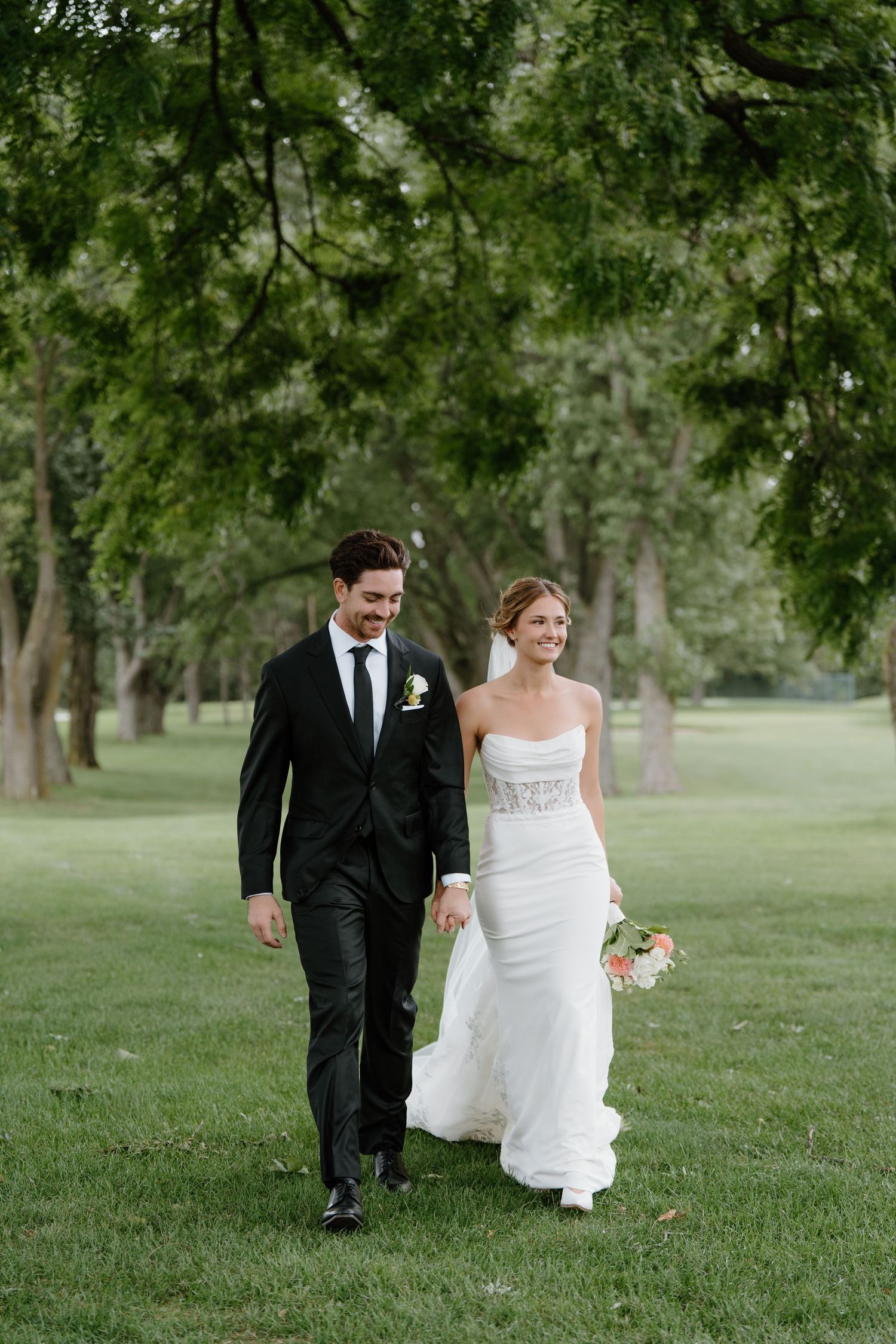 A bride and groom walk together across a lush green lawn surrounded by trees on their wedding day.