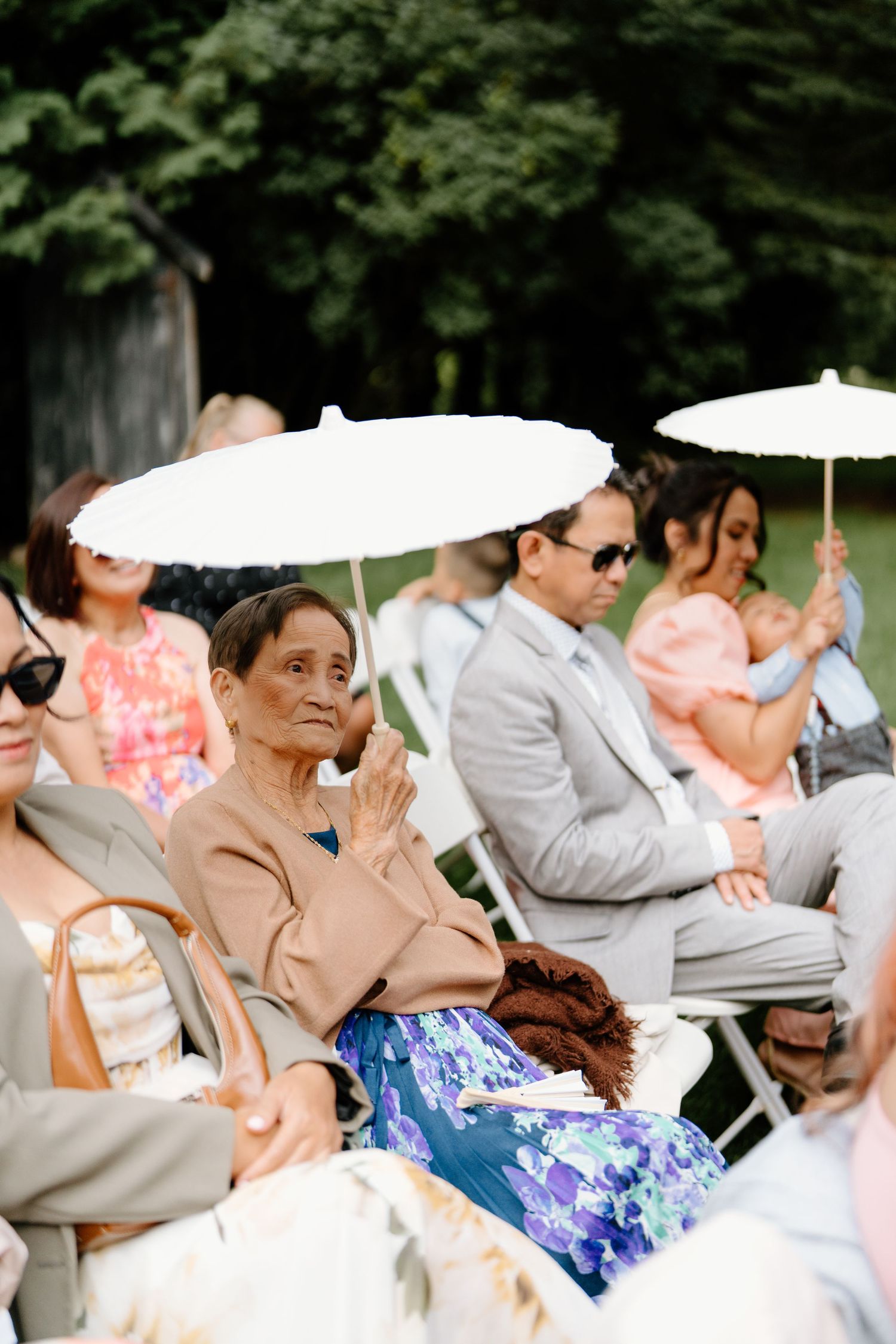 Wedding guests seated in white chairs outdoors react with emotion during ceremony.