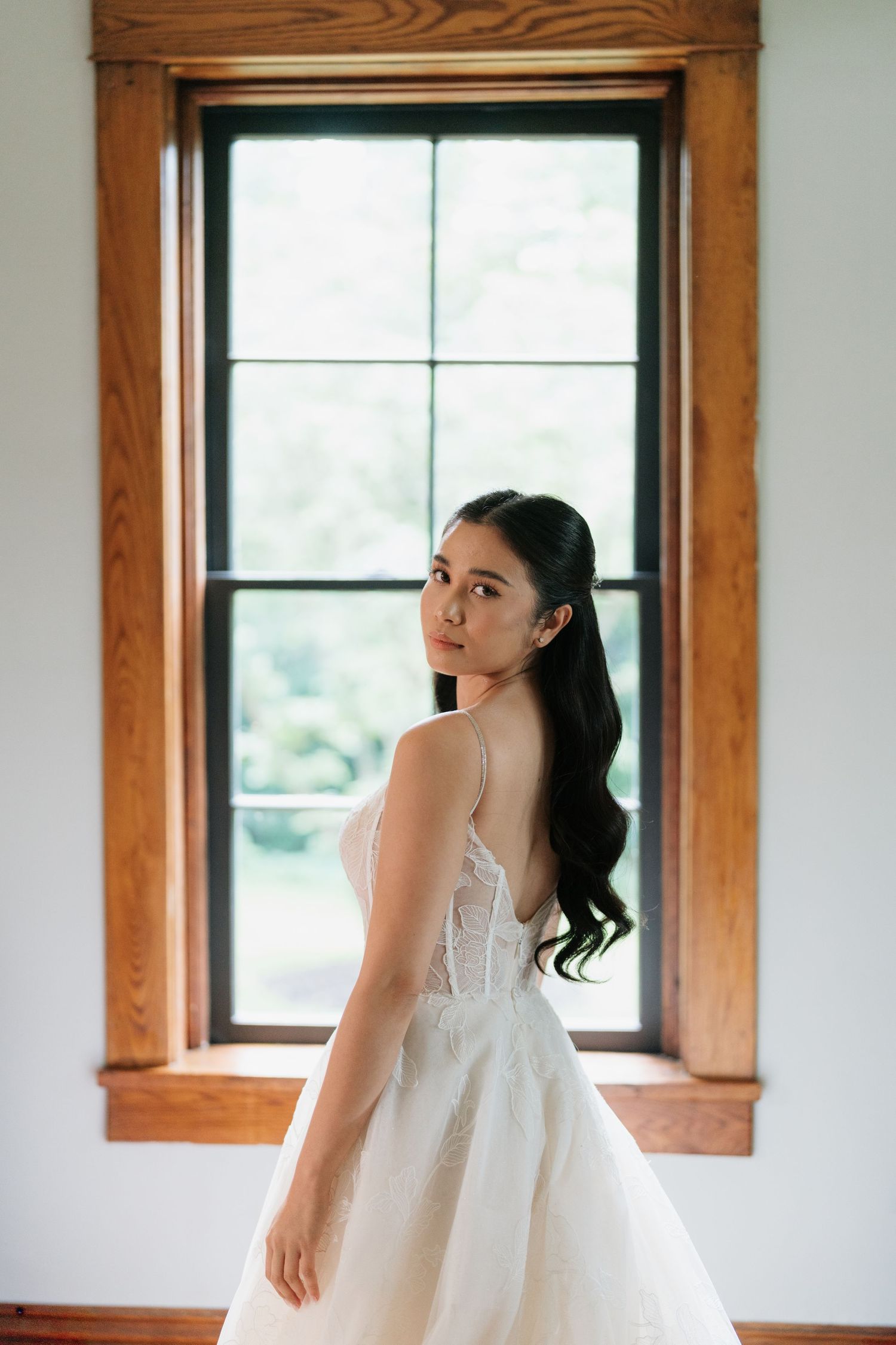 A bride in a white lace wedding dress twirls gracefully near sunlit windows with wooden trim.