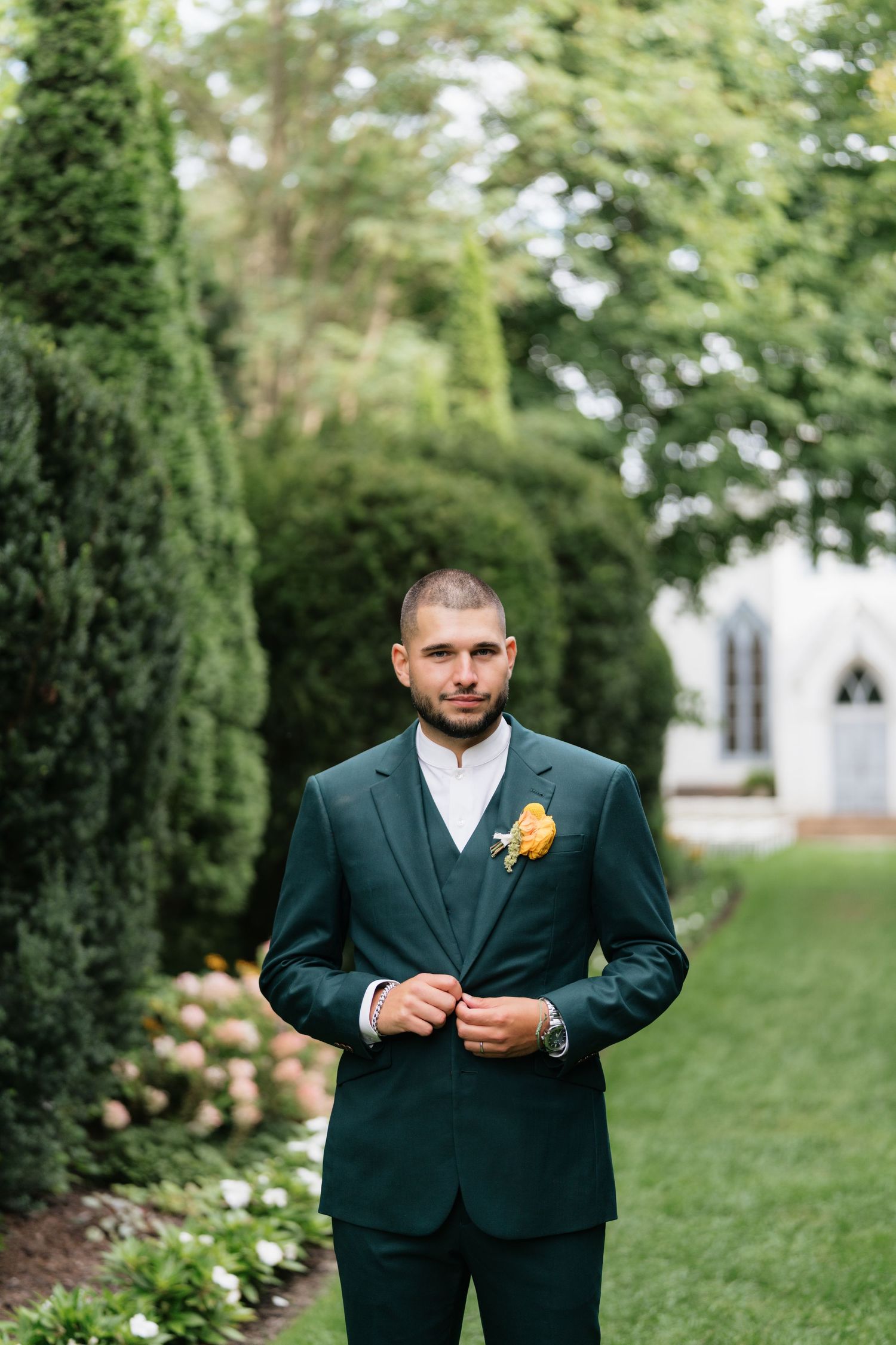 A person in a dark suit adjusts their boutonniere while standing on a grassy lawn with trees in the background.