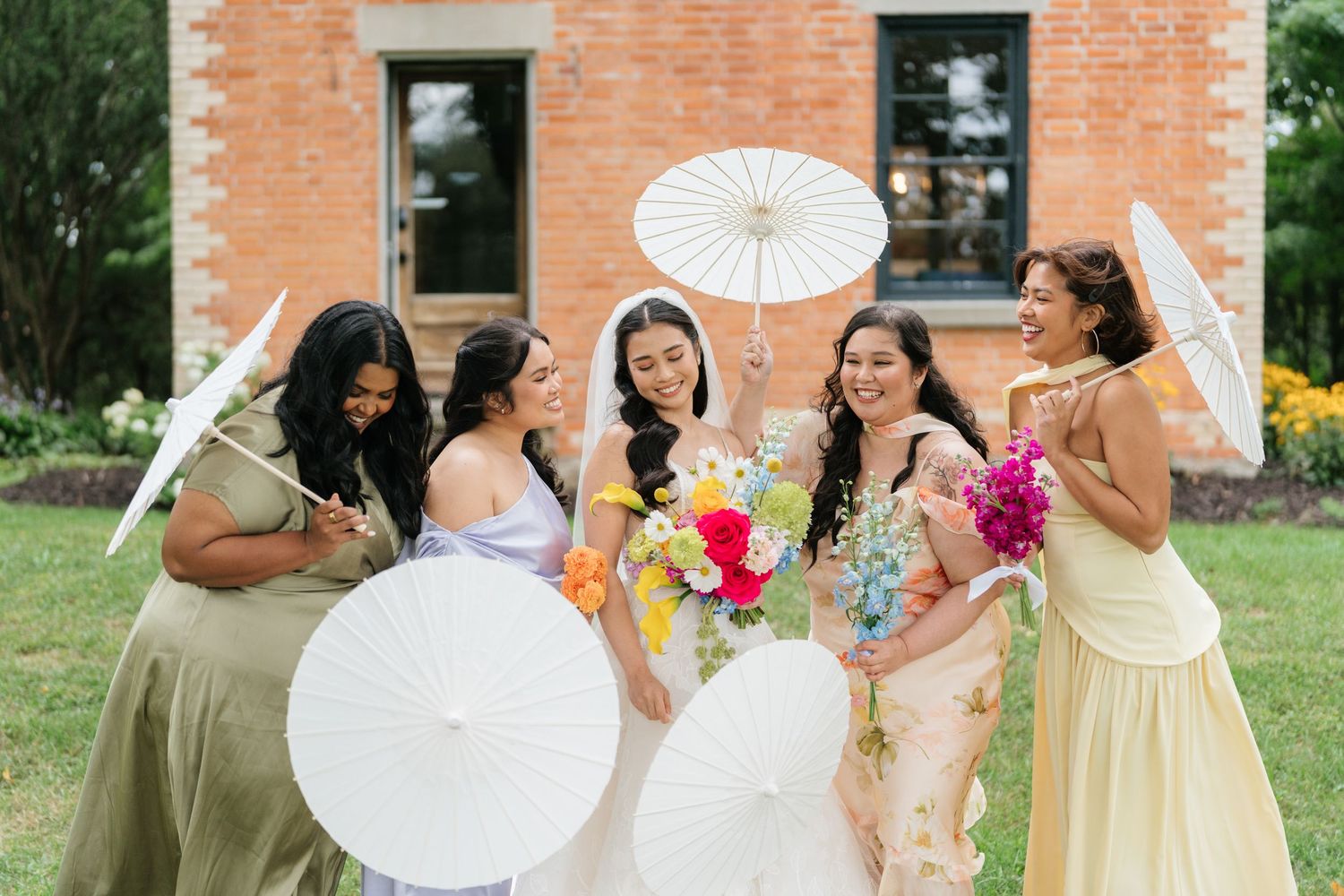 A bridal party in pastel dresses shares joyful laughter while holding white paper parasols on a sunny day.