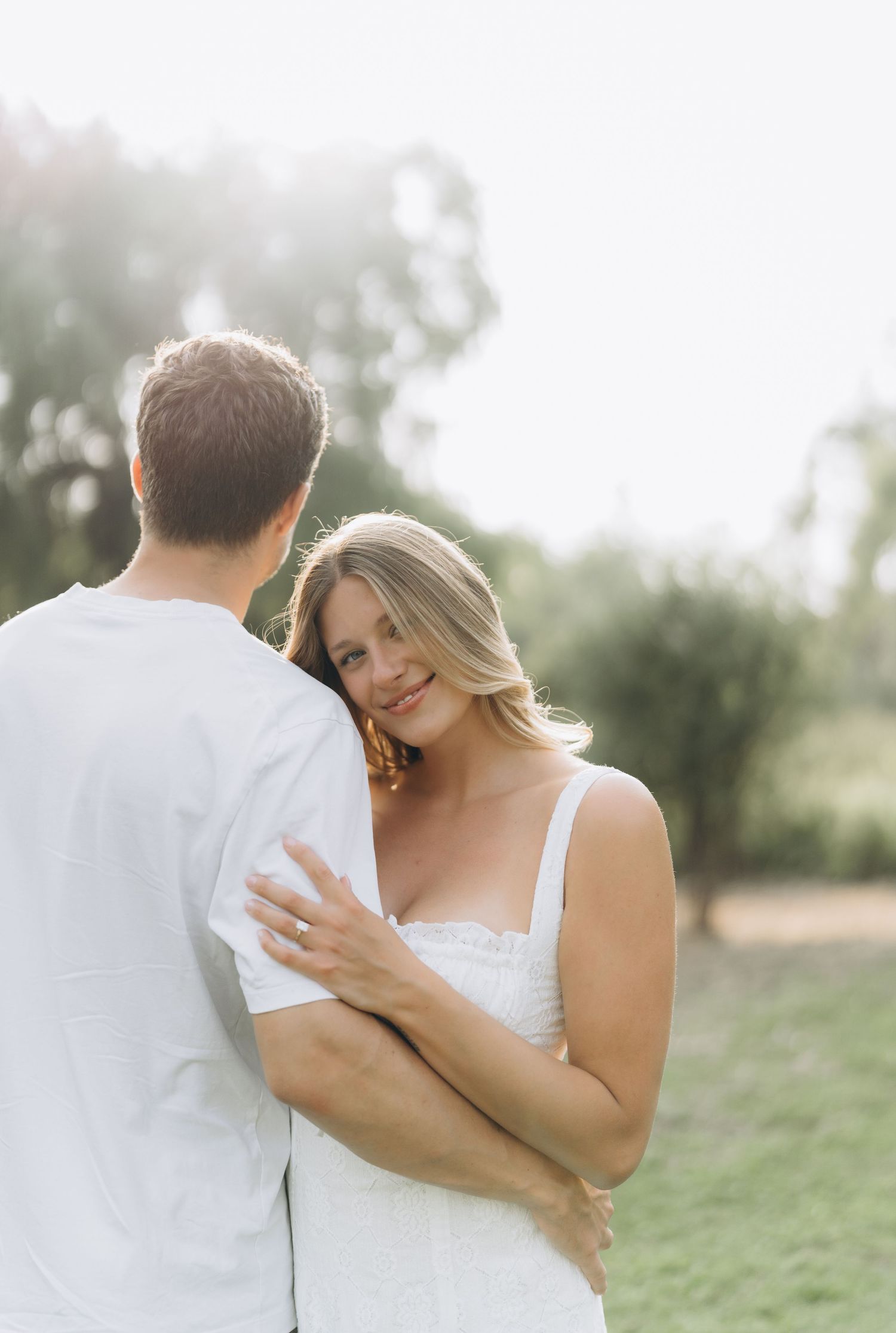 A romantic couple embraces outdoors in white attire during a wedding photoshoot with soft natural lighting.