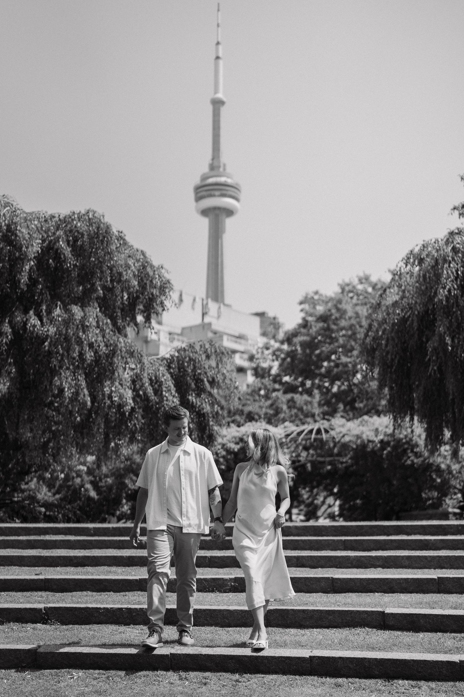 A couple walks up stone steps in a park with Toronto's CN Tower visible in the black and white background.