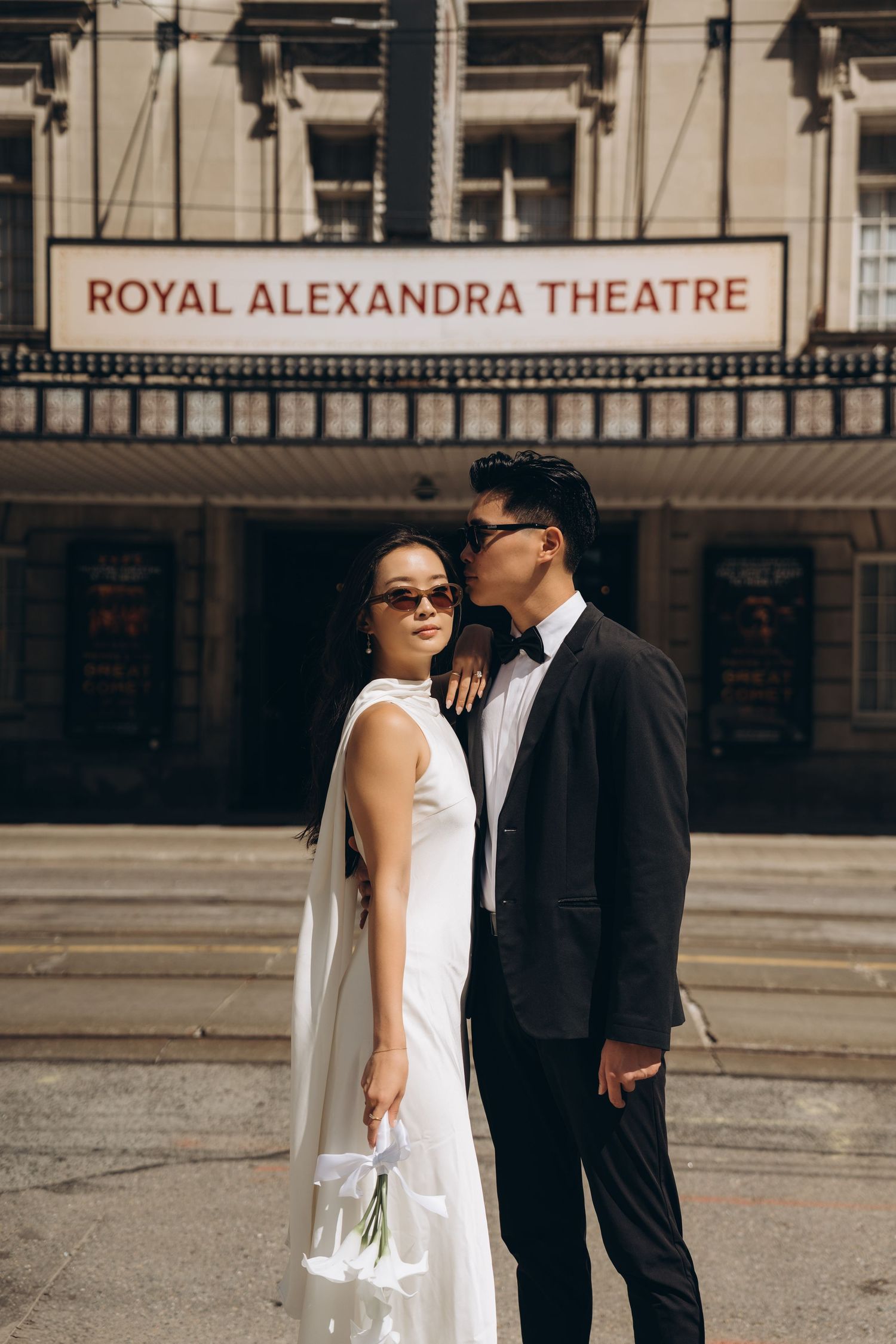 Couple in wedding attire poses outside Royal Alexandra Theatre with bride in white dress and groom in black suit.