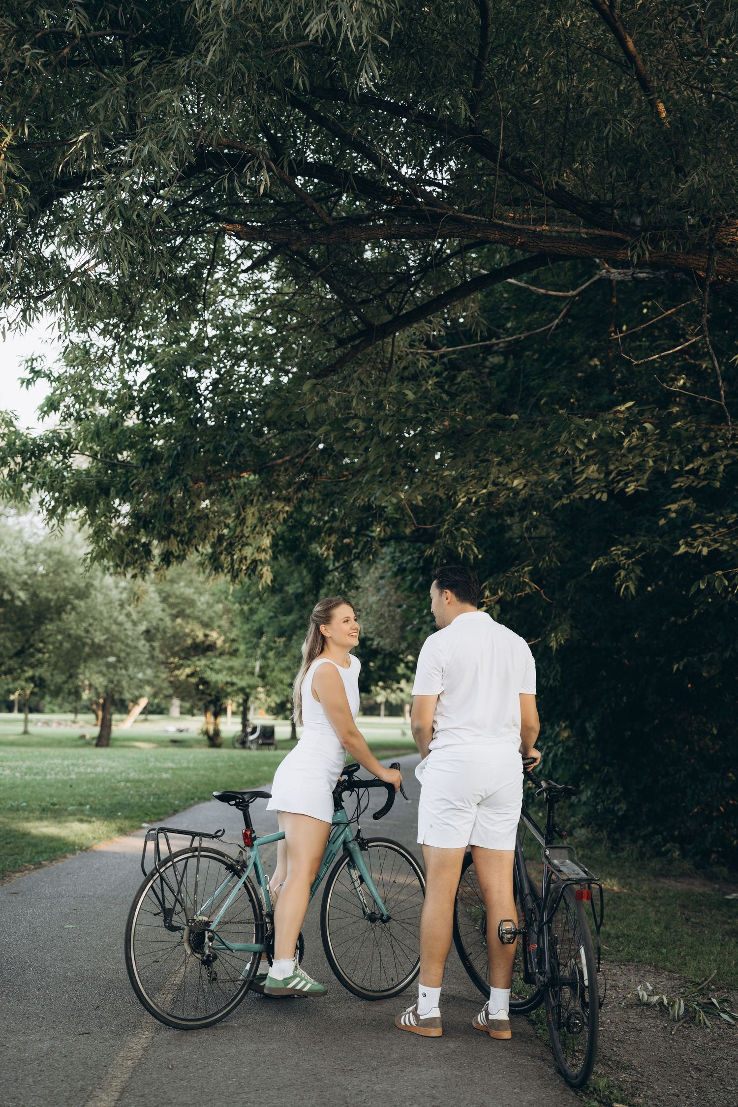 Two people in white outfits stand with their bicycles on a park path under shady trees.