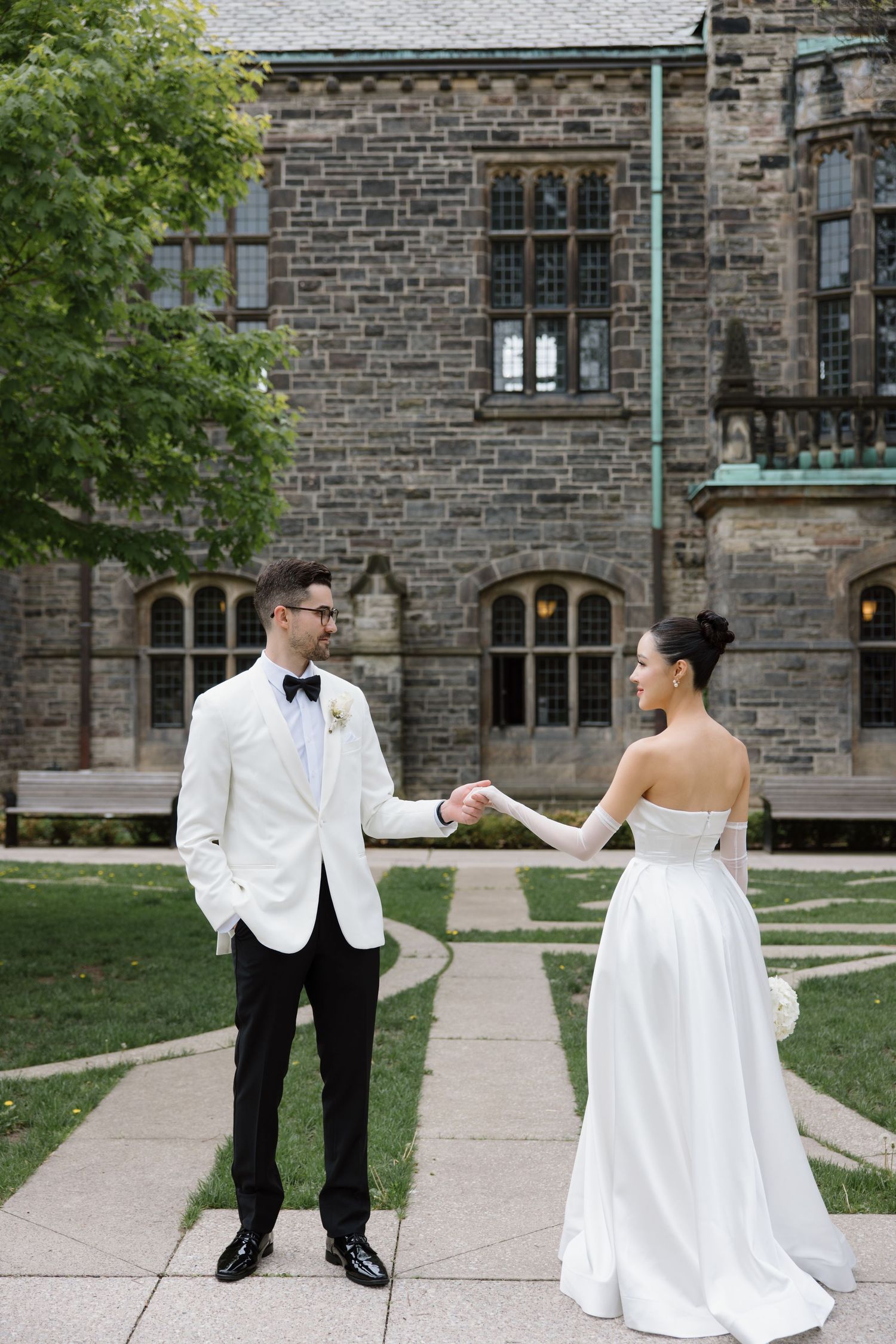 A couple in elegant wedding attire holding hands on a stone path outside a historic Gothic-style building.