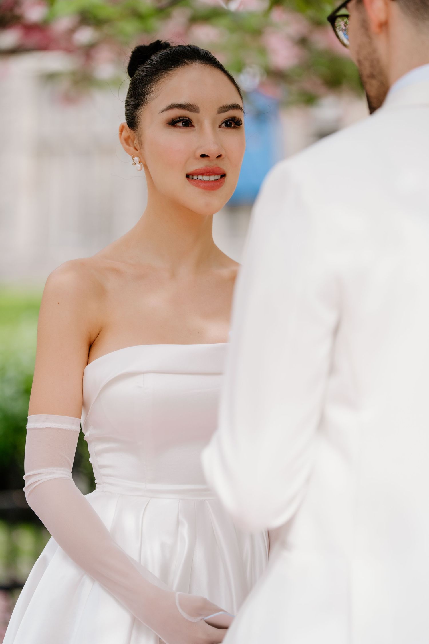 Elegant bride in a white strapless gown with sheer gloves gazes lovingly during a spring wedding ceremony outdoors.