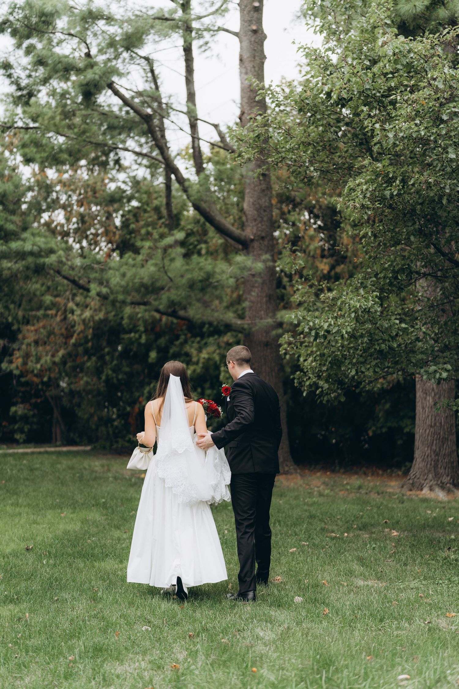 Newlyweds walking together on a grassy lawn surrounded by tall trees in a romantic outdoor setting.