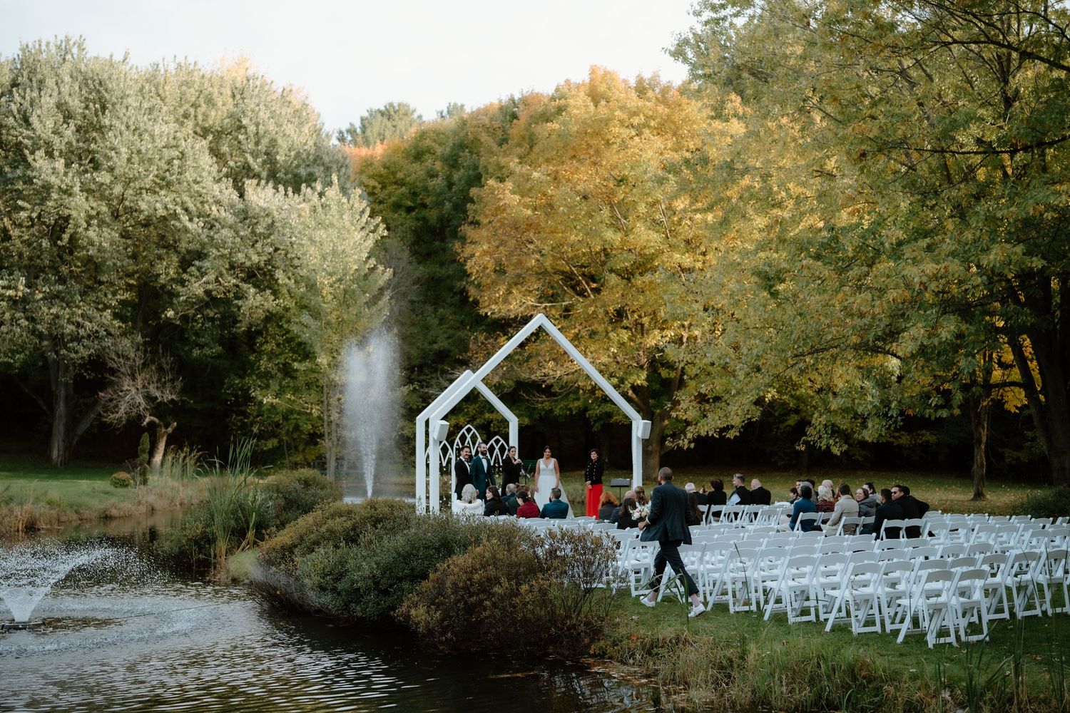 Outdoor wedding ceremony site with white chairs and garden arch near a pond with autumn trees.