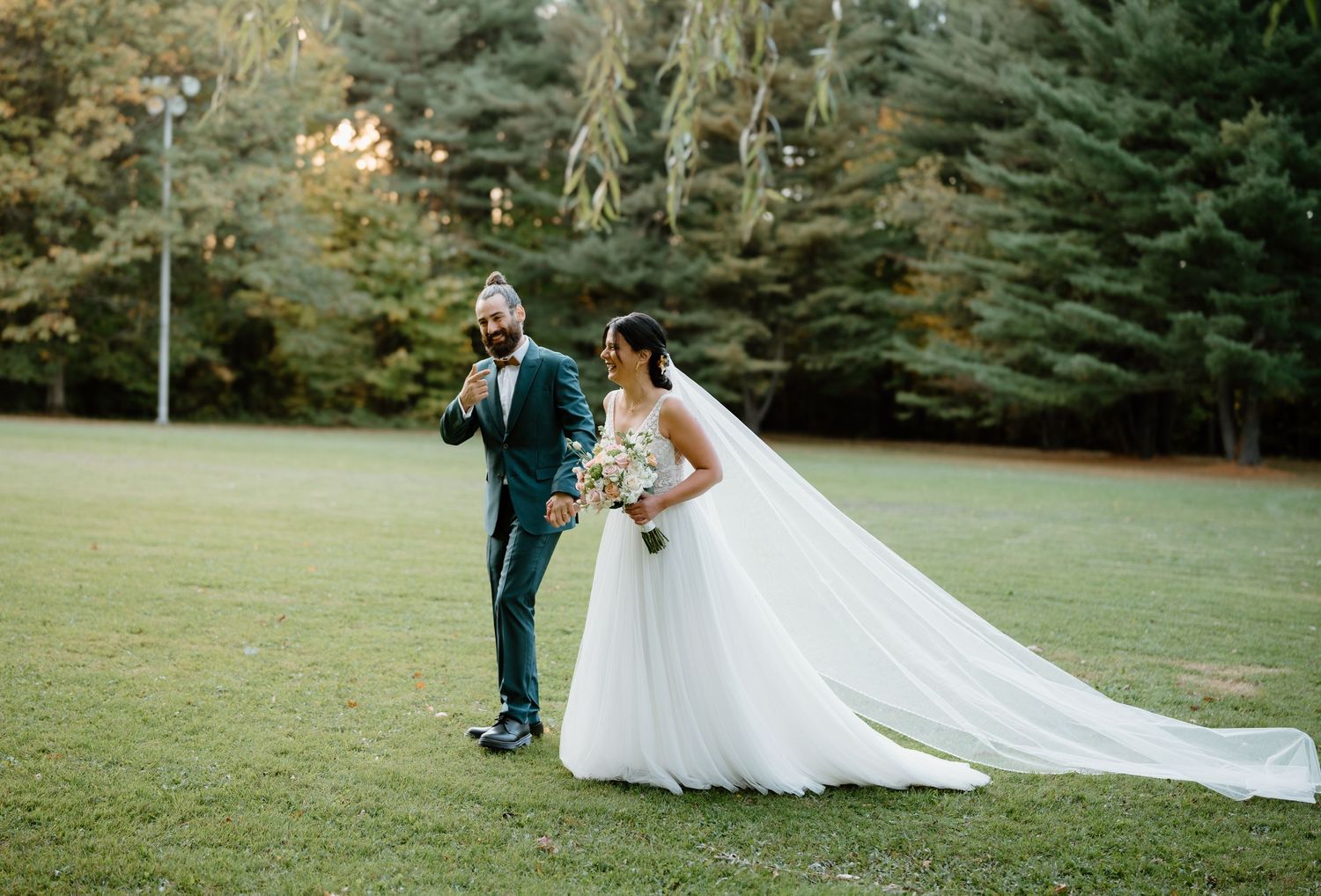 A bride in flowing white gown and groom in teal suit pose outdoors against evergreen trees.