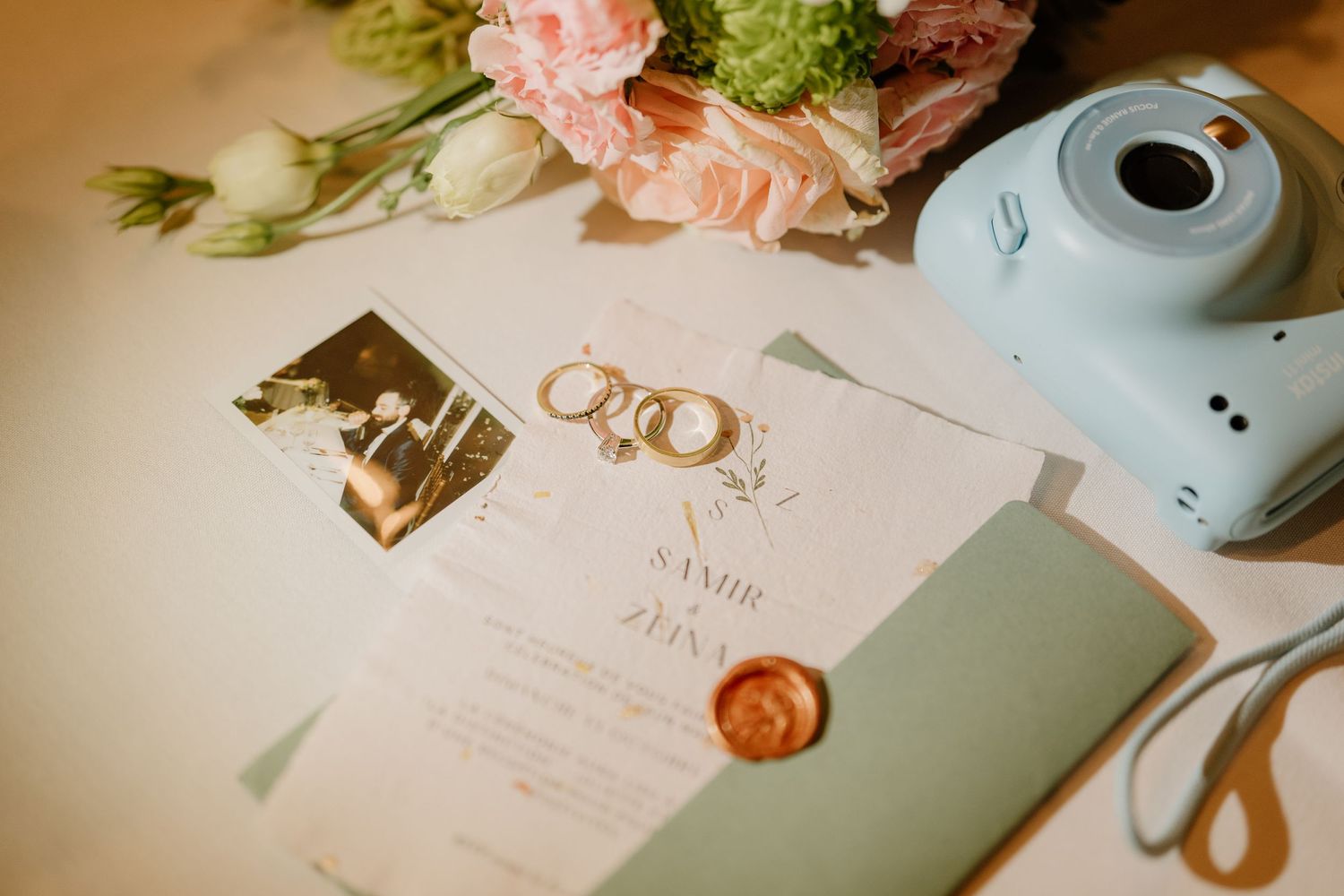Wedding stationery with wax seal, rings, and a mint blue Instax camera on white surface.
